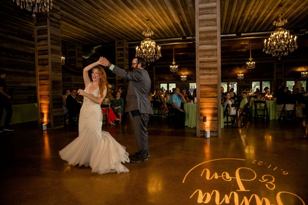 A couple dances together in a rustic venue with wooden walls, chandeliers, and a light-up floor design.