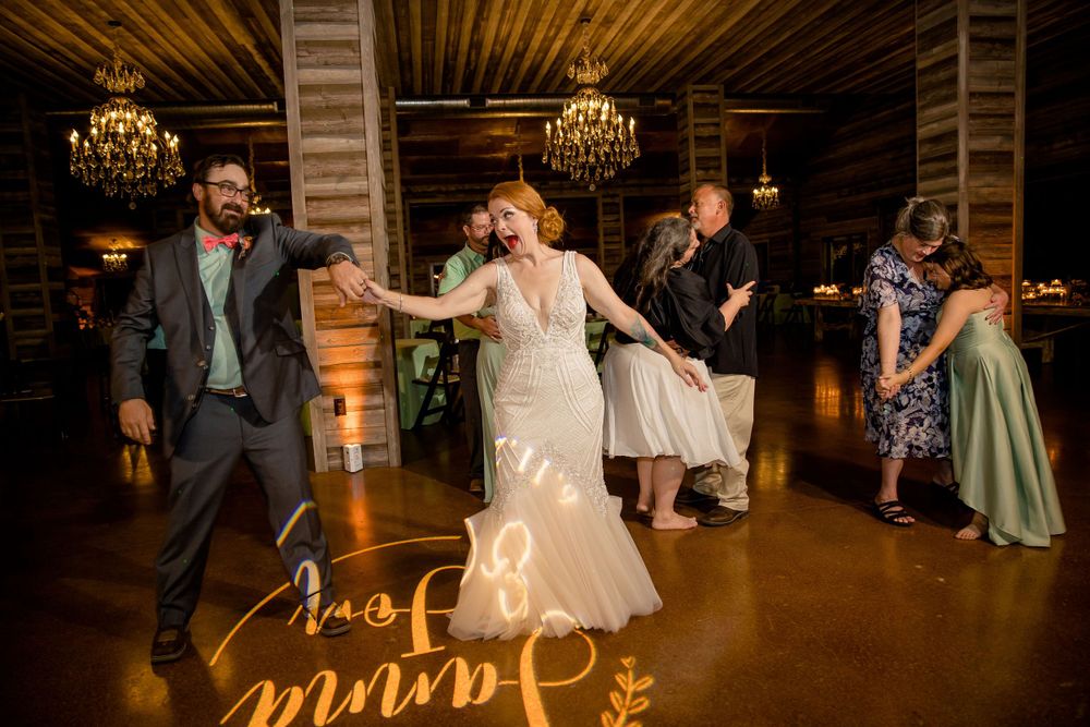 Wedding party dancing energetically in a rustic venue with wooden beams and elegant chandeliers.