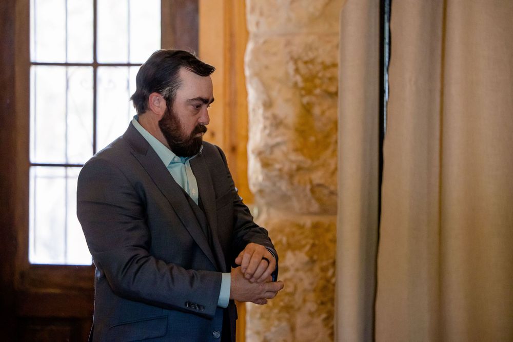 Man in formal attire adjusting his sleeve by a window.
