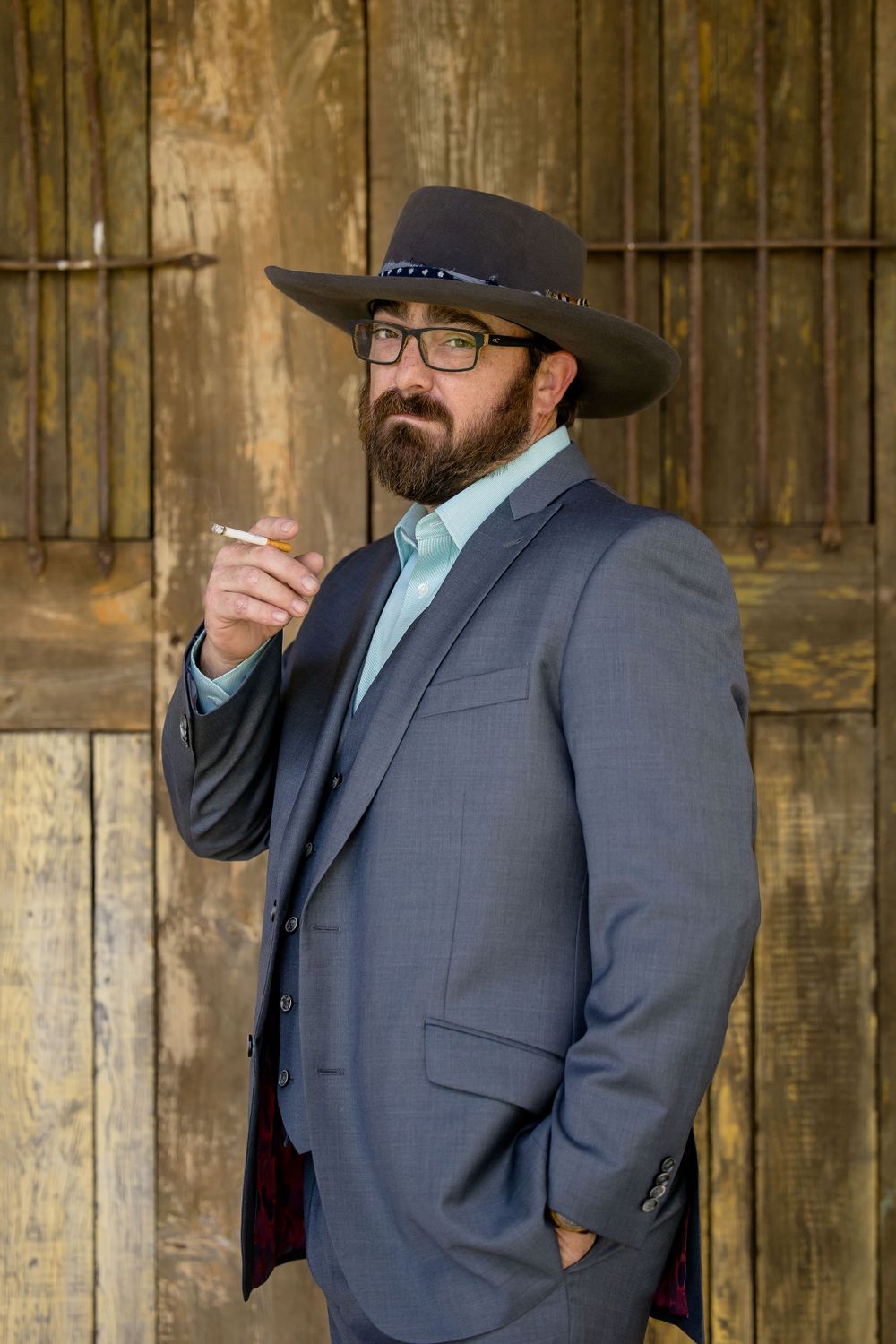 Man in grey suit and western hat posing against rustic wooden wall.