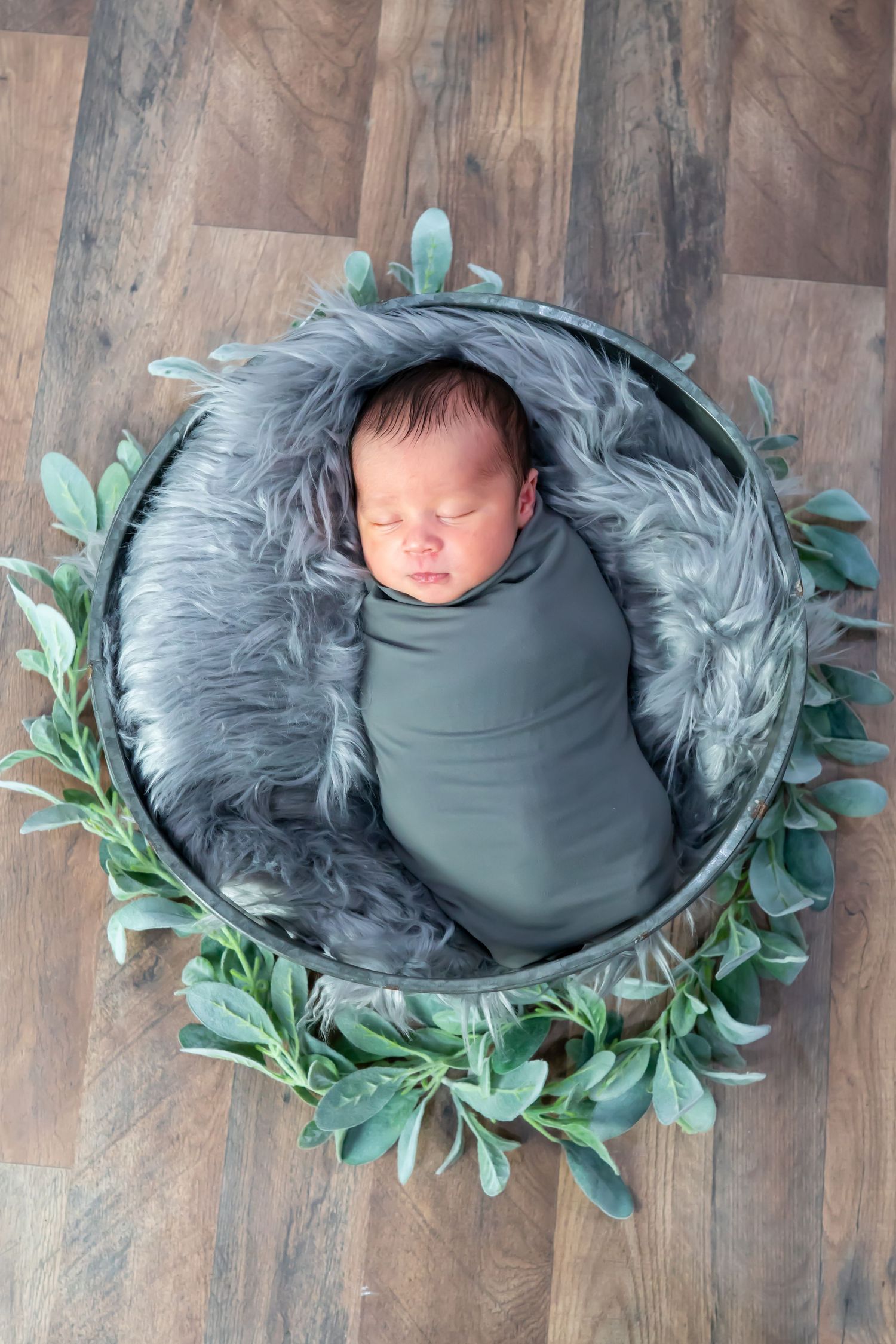 Peacefully sleeping newborn swaddled in gray wrap, nestled in a round bowl with fur lining and greenery.