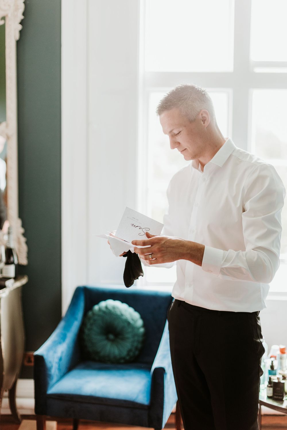 Person in white shirt reading a card while standing next to a teal velvet chair.