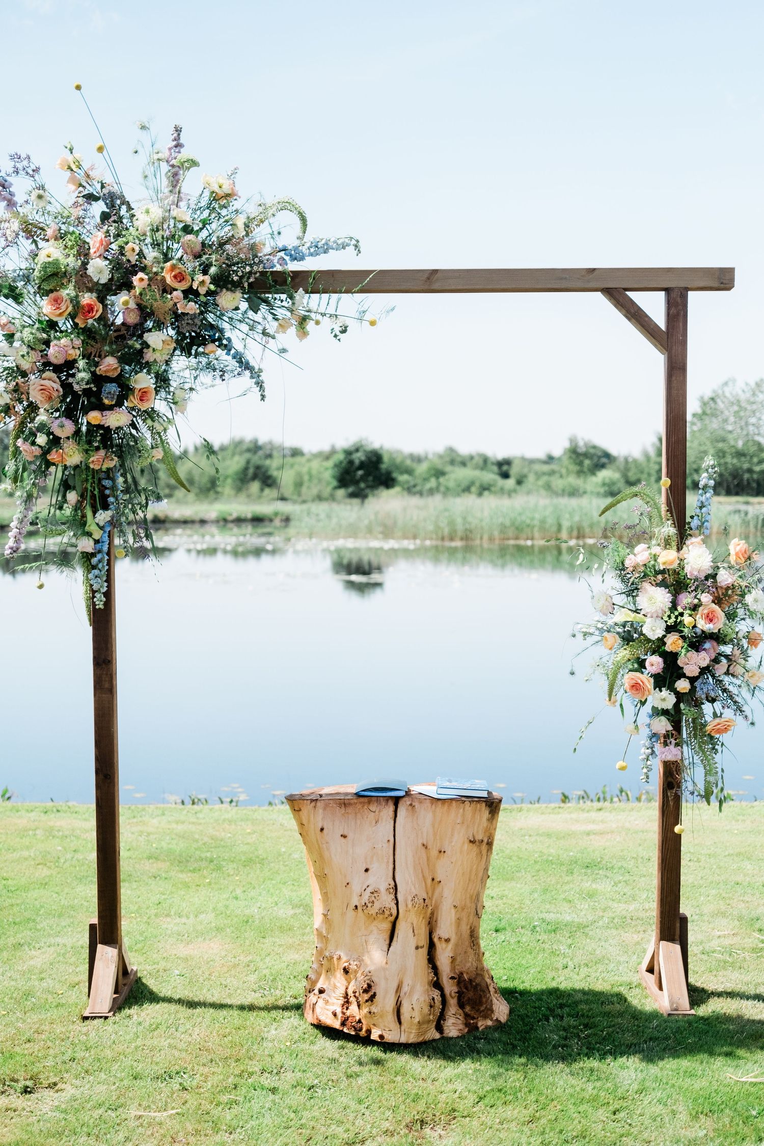 A wooden wedding arch decorated with peach and white flowers overlooks a serene lake.