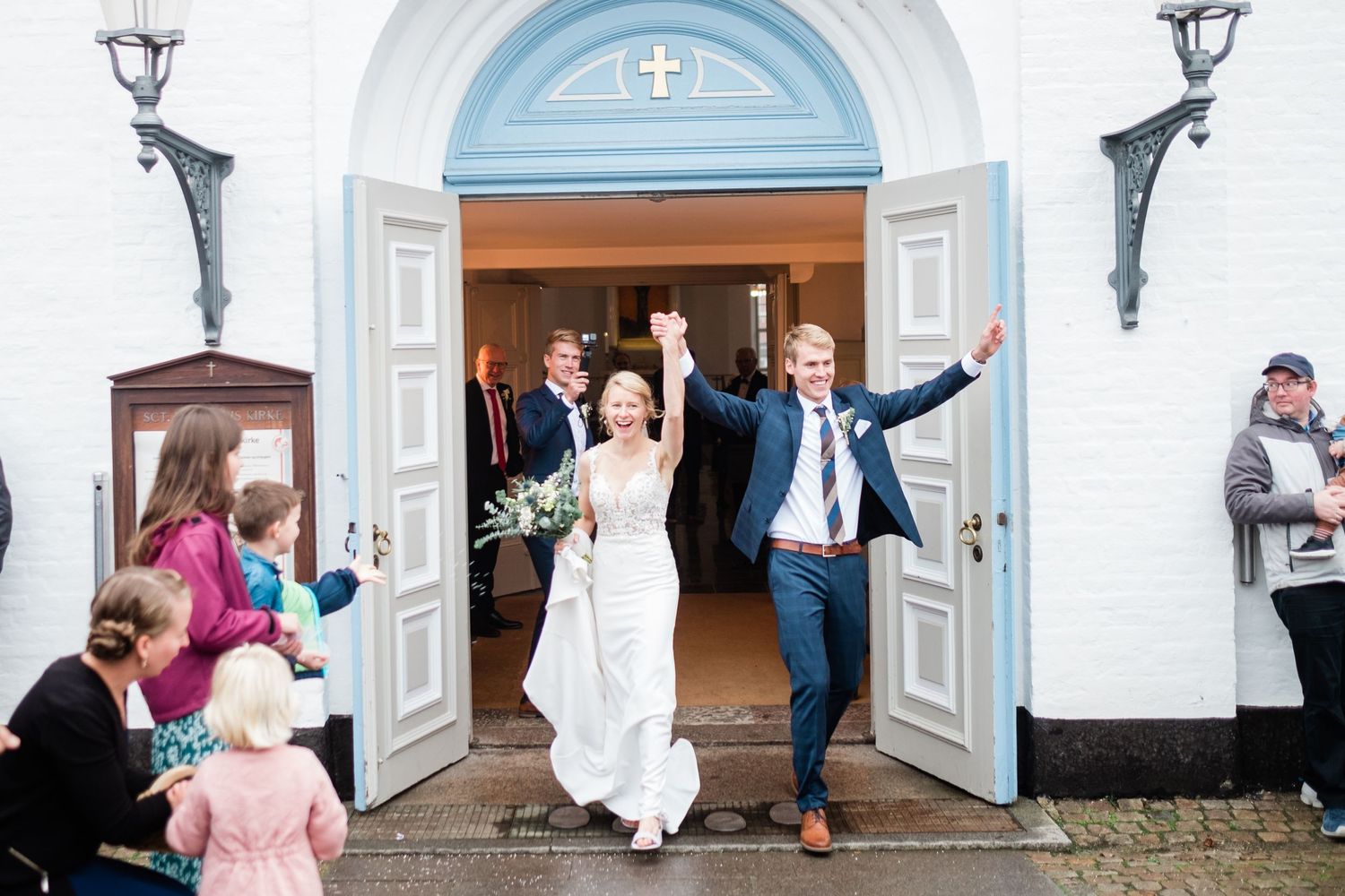 Newlyweds exit church with raised arms while wedding guests celebrate with confetti.