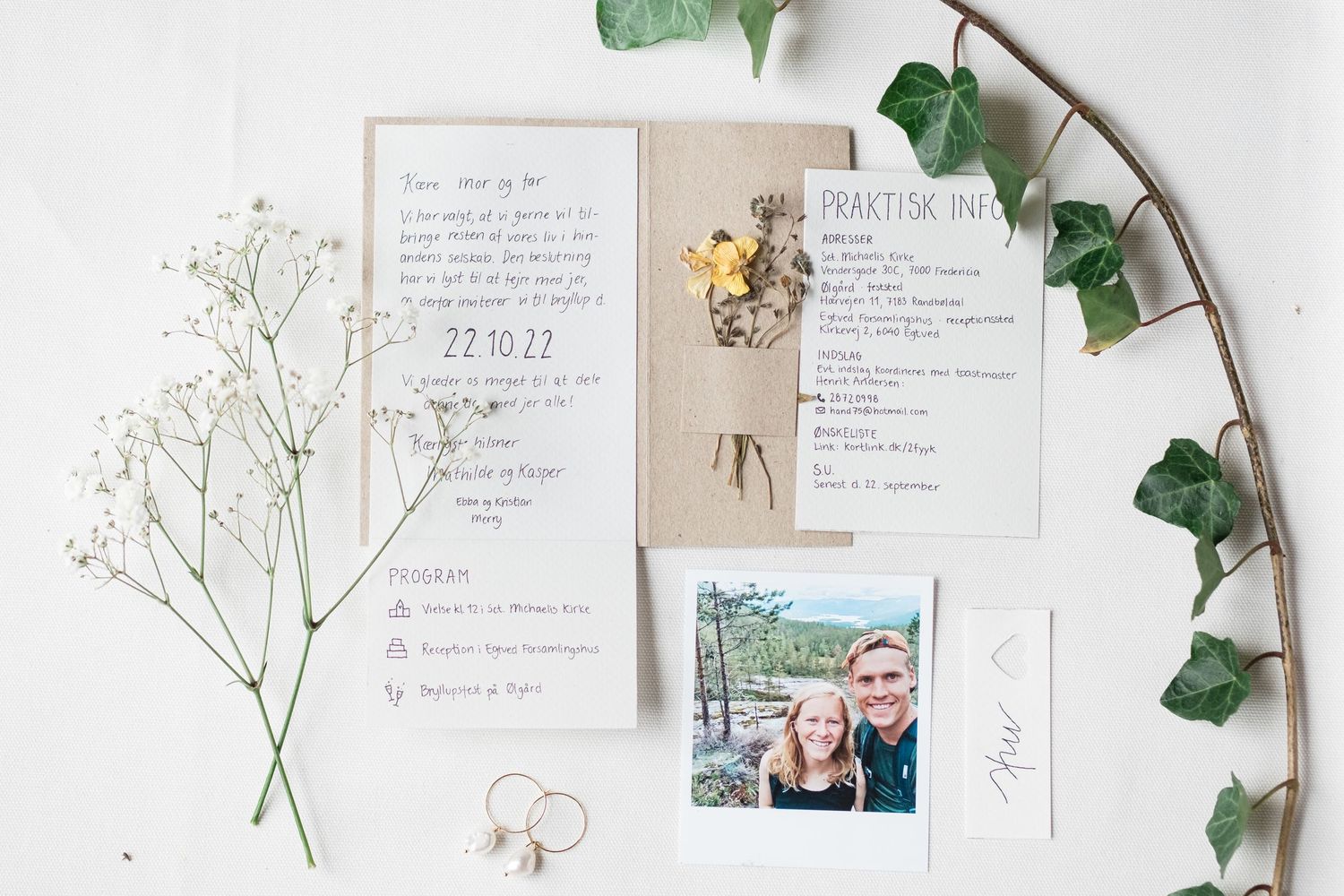 Wedding stationery flat lay with invitations, rings, and greenery on white background.