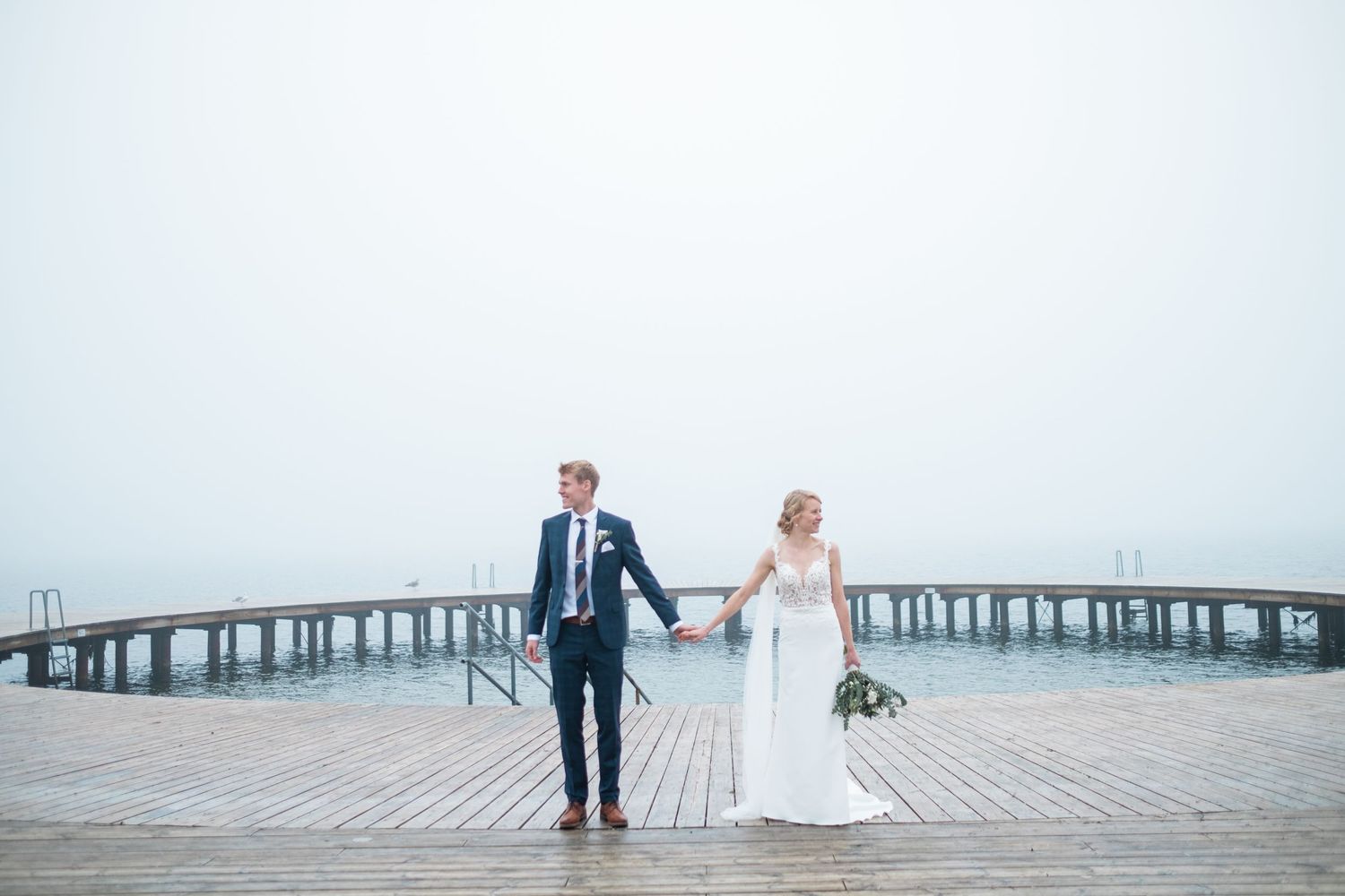 A wedding couple stands together on a wooden pier overlooking a misty ocean landscape.