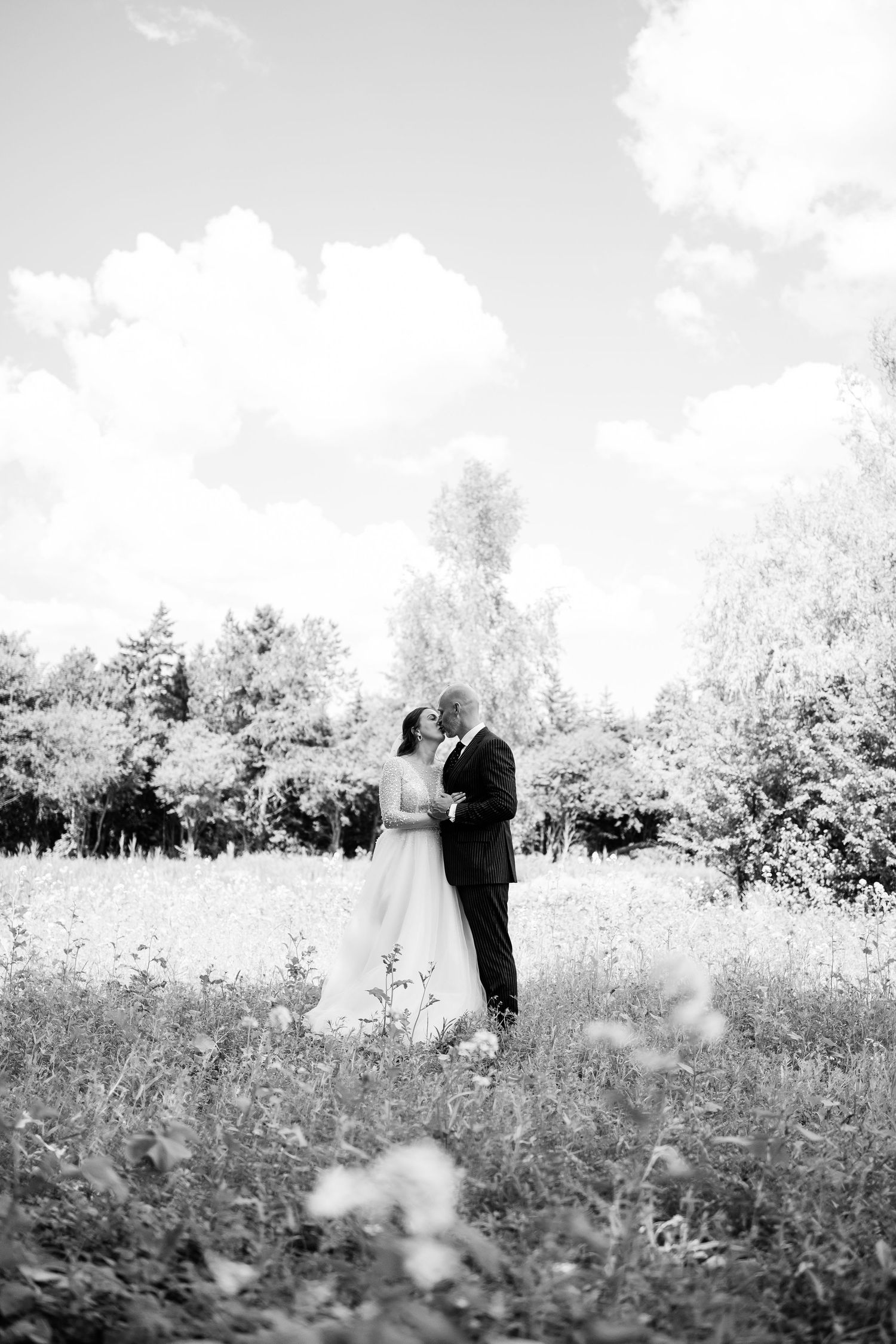 A black and white portrait of a couple embracing in a meadow with puffy clouds overhead.