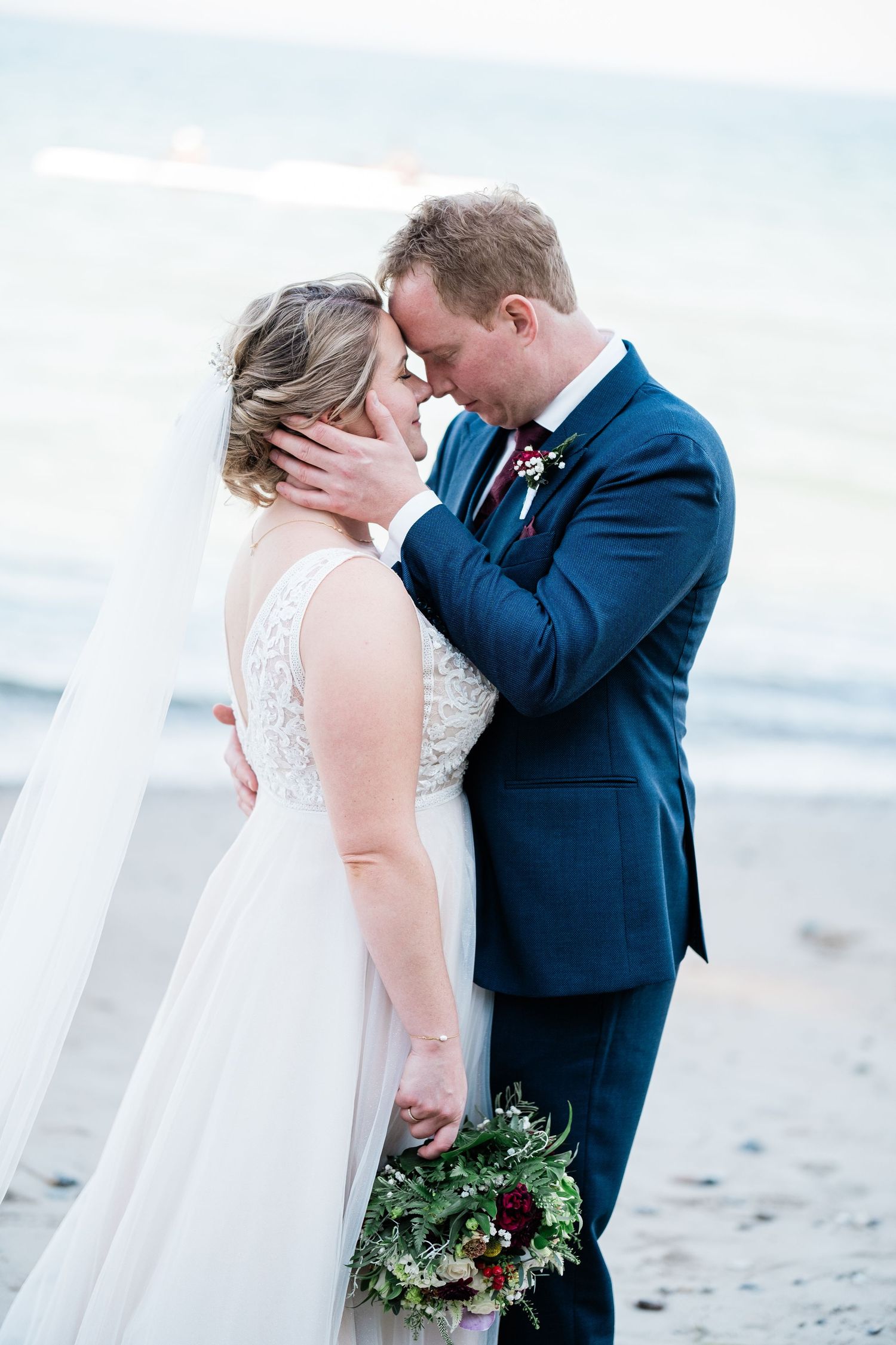 A couple shares an intimate moment on a beach, with the bride holding a bouquet and wearing a flowing white dress.