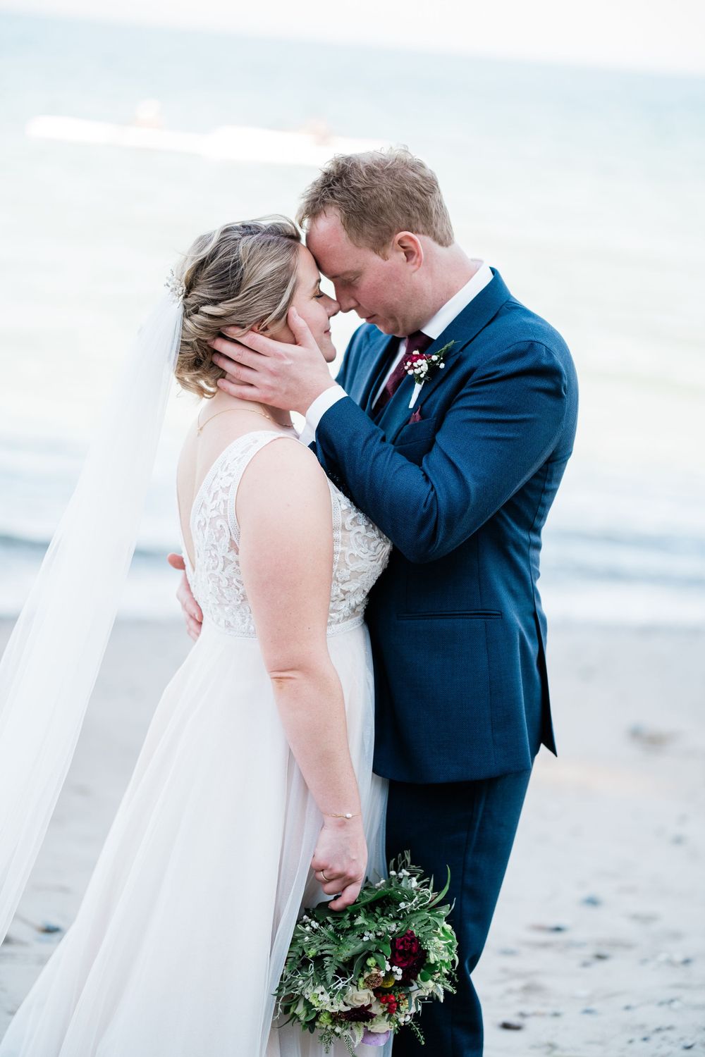 A couple shares an intimate moment on a beach, with the bride holding a bouquet and wearing a flowing white dress.