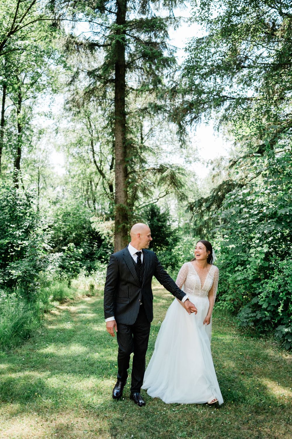 A couple in wedding attire stands together on a sunlit forest path surrounded by lush green trees.