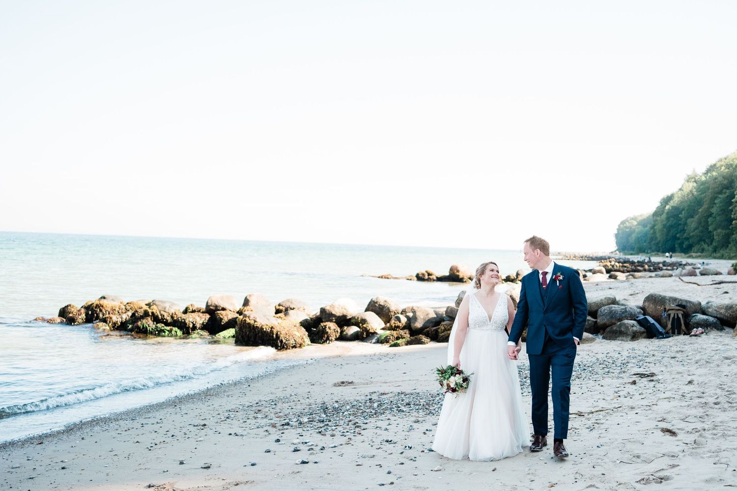 A couple walks along a rocky beach shoreline in formal wedding attire.