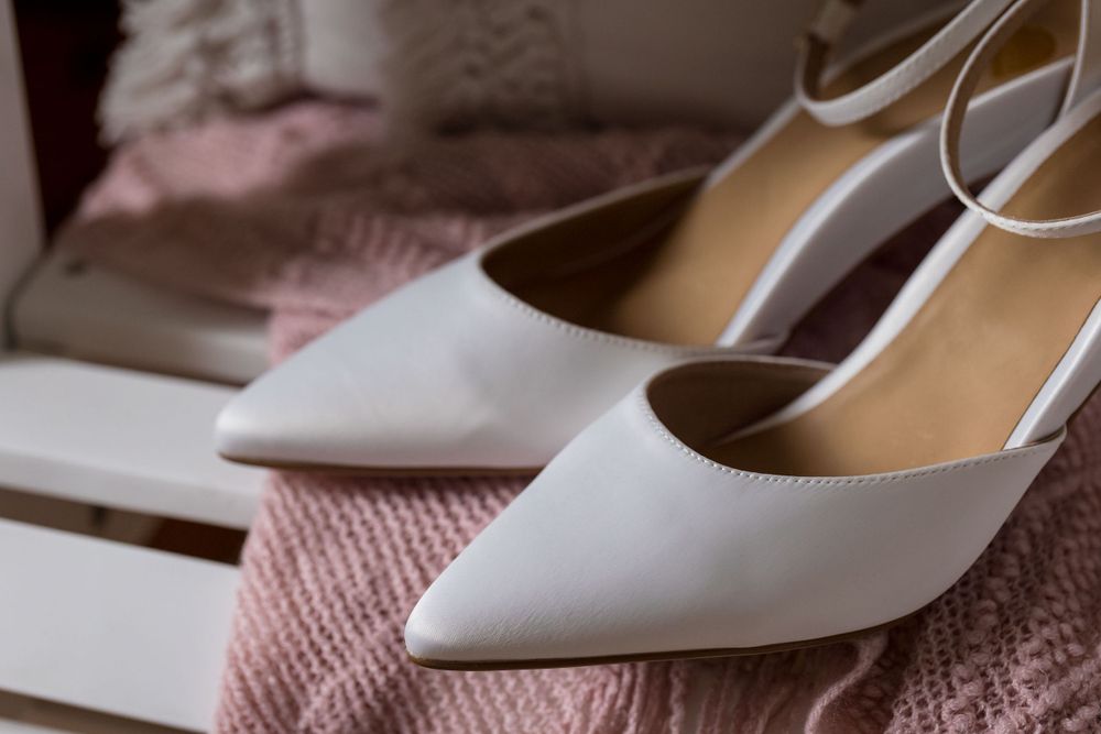 Elegant white pointed-toe bridal shoes with ankle straps rest on pink fabric.