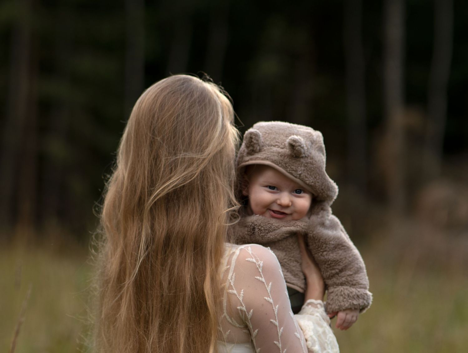 En långhårig brunnett kvinna bakifrån håller om sin bebis som ler mot kameran. Bebisen har en ljusbrun luvdräkt på sig.