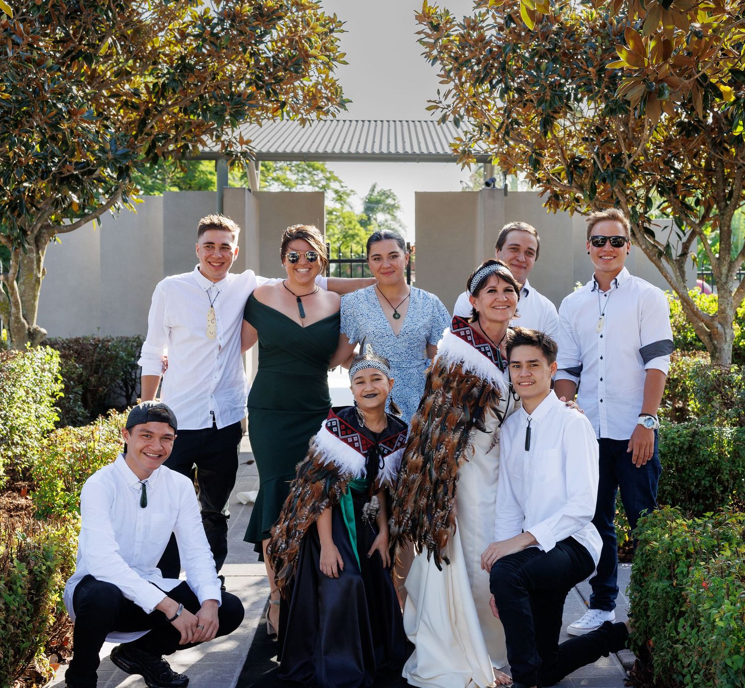 Wedding party poses together outdoors in formal attire against a garden backdrop.