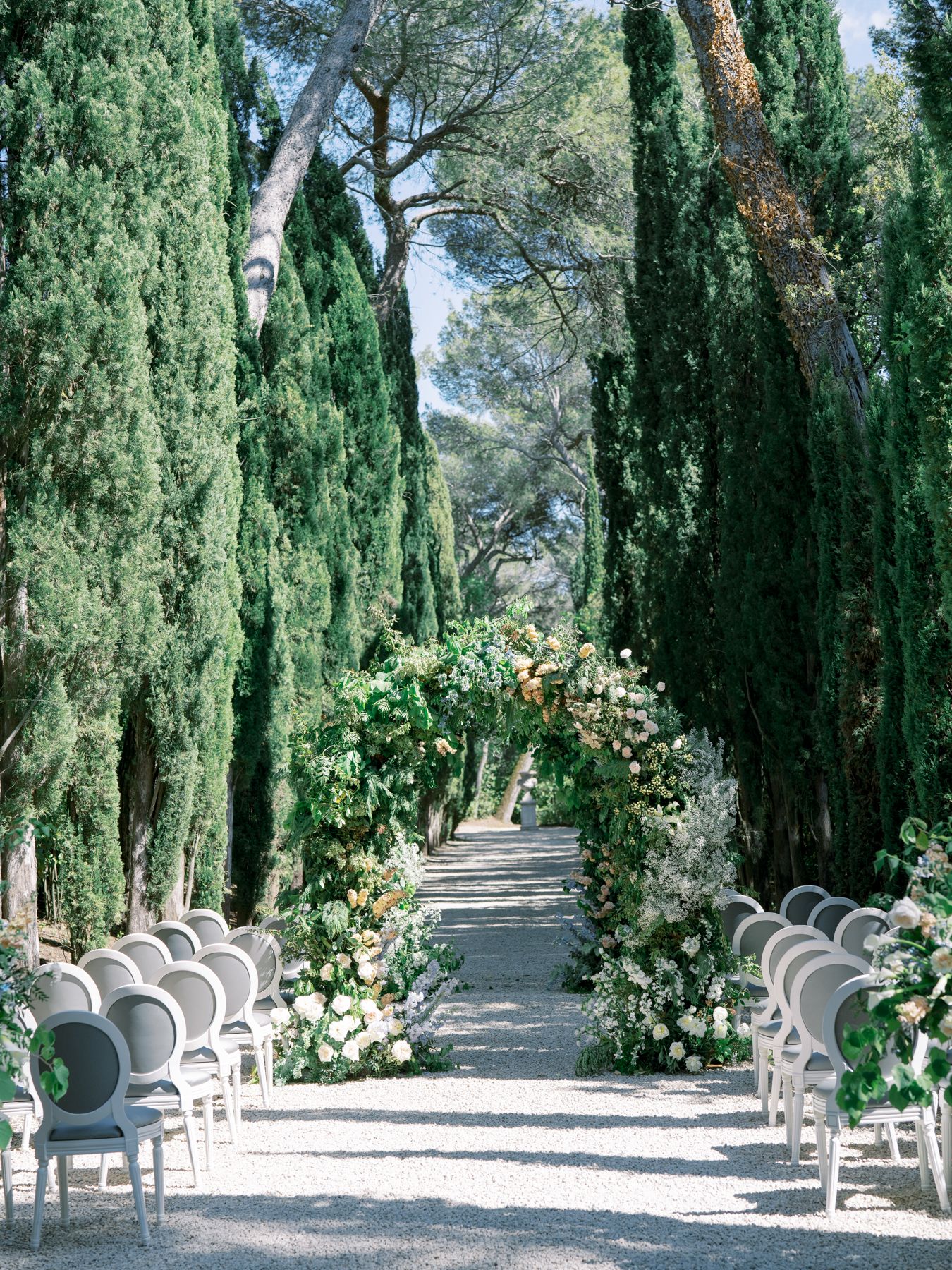 An elegant outdoor wedding ceremony setup with rows of chairs along a cypress tree-lined path.