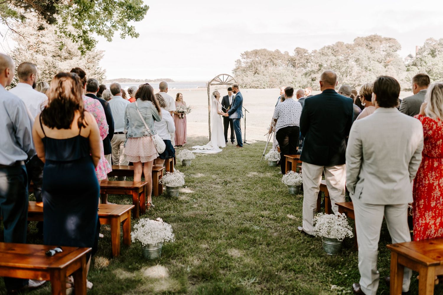 Wedding guests gather on wooden benches in a garden setting with natural decor.