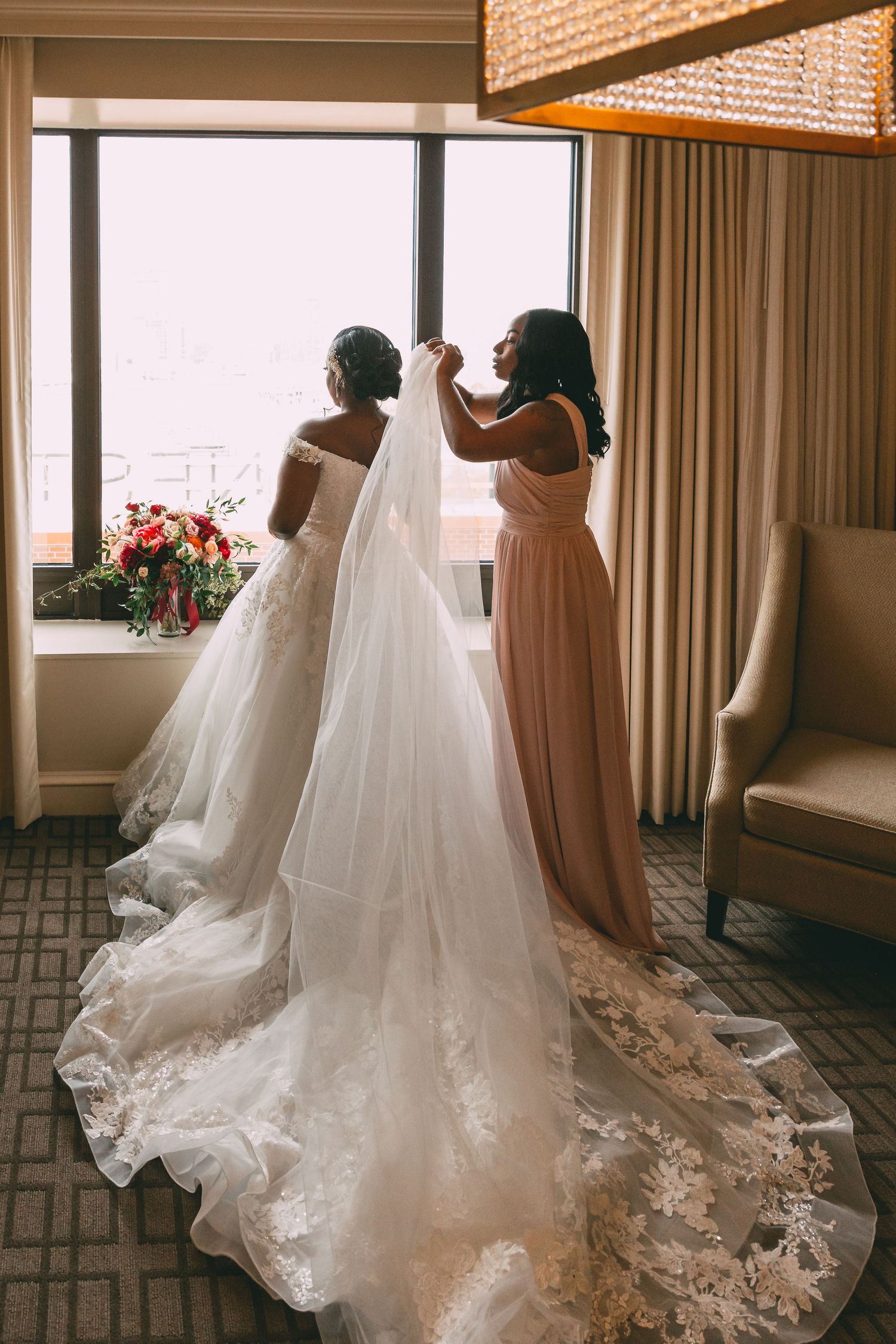 Bride in elegant white gown with lace train gets veil adjusted by bridesmaid in blush dress near hotel window.
