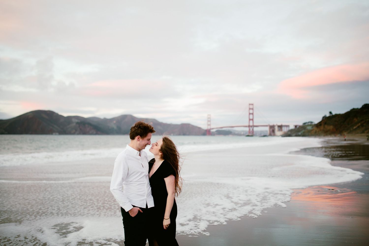Couple shares romantic moment at Baker Beach with Golden Gate Bridge in background at sunset.