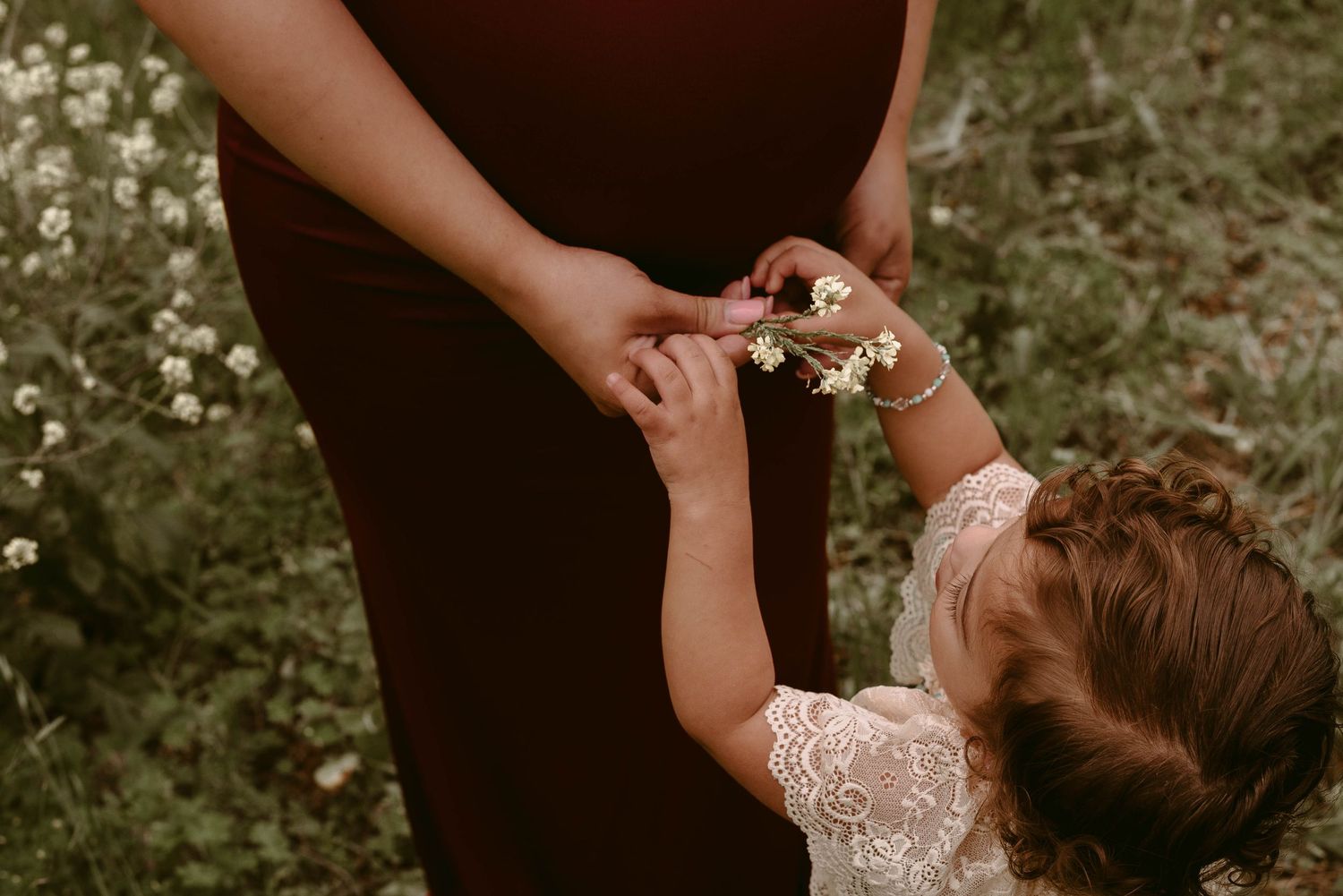 Small child's hand reaches up to touch a pregnant belly in a touching outdoor moment.