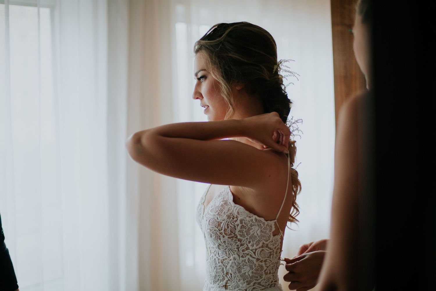 A bride in a lace dress adjusts her hair while getting ready by a window in soft natural light.