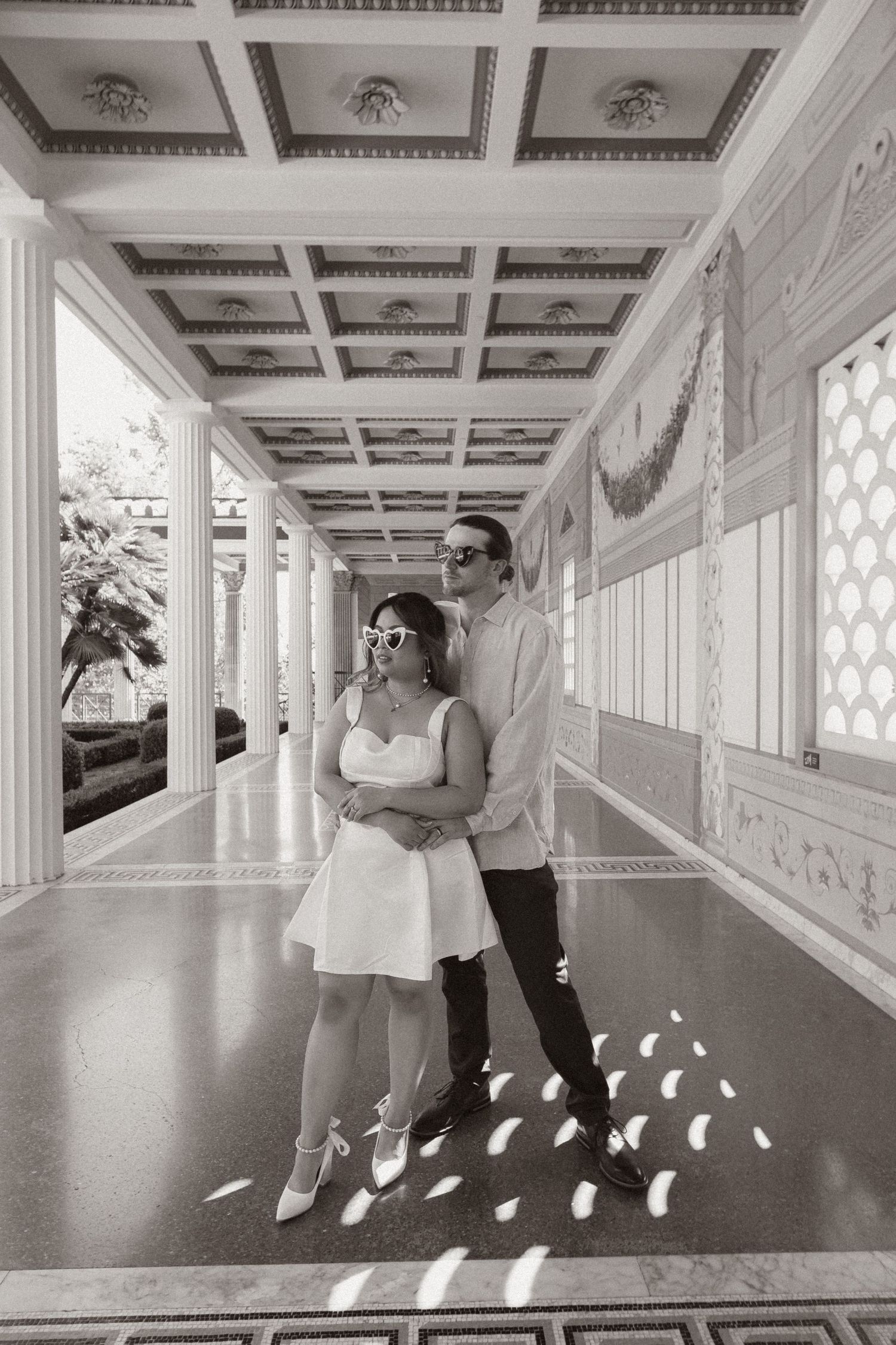 A couple poses in an elegant corridor with ornate white coffered ceiling and patterned light.