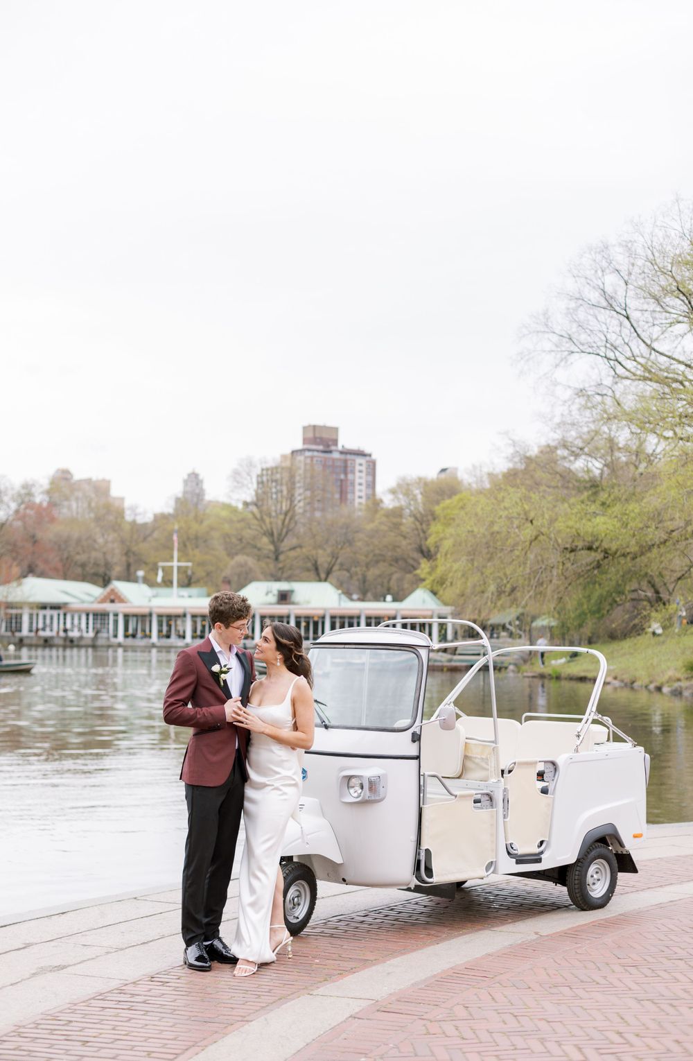 A couple poses with a white vintage cart by a lake with city buildings in the background.