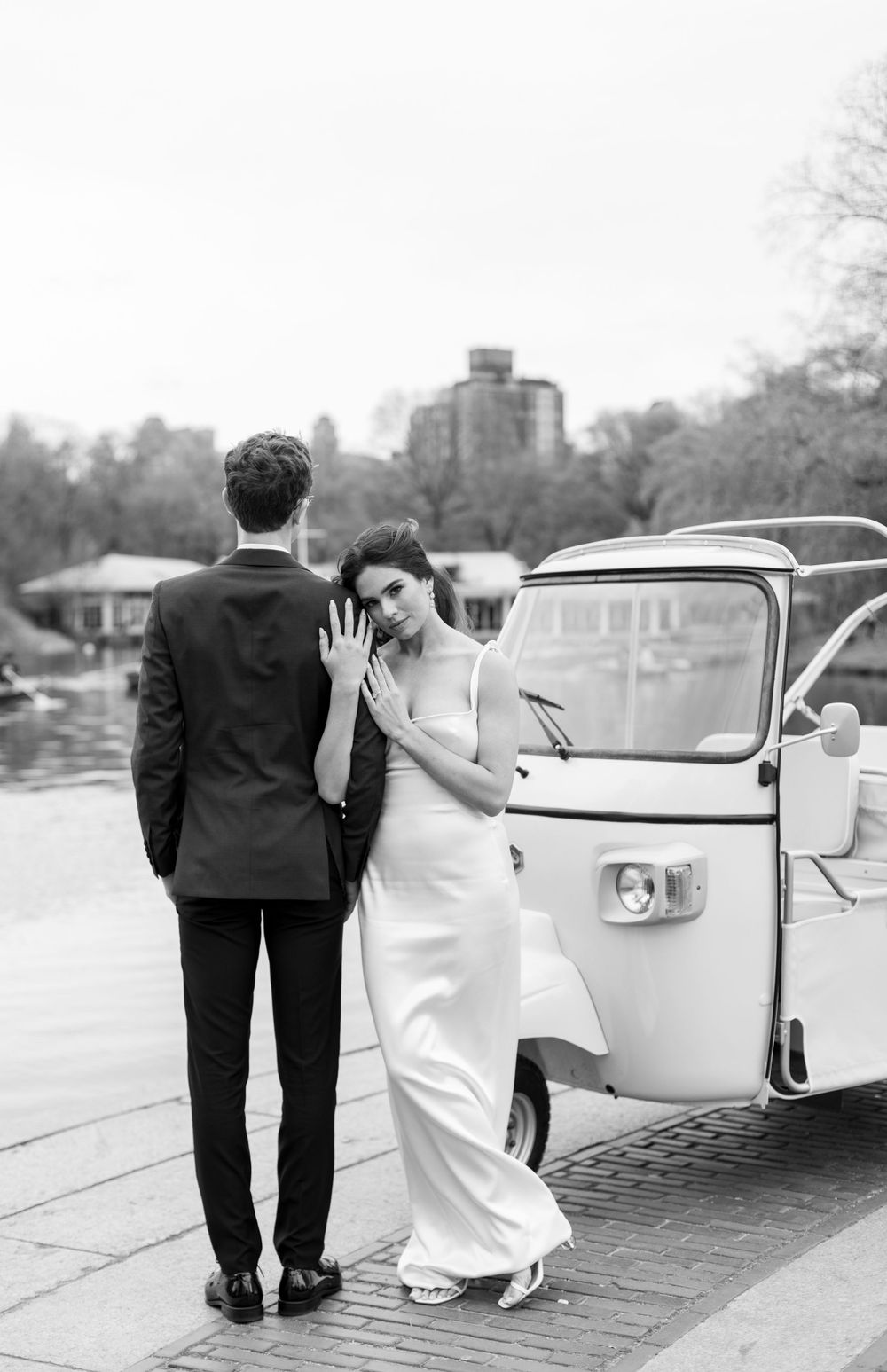A black and white photograph of a couple near a vintage vehicle during their wedding day.