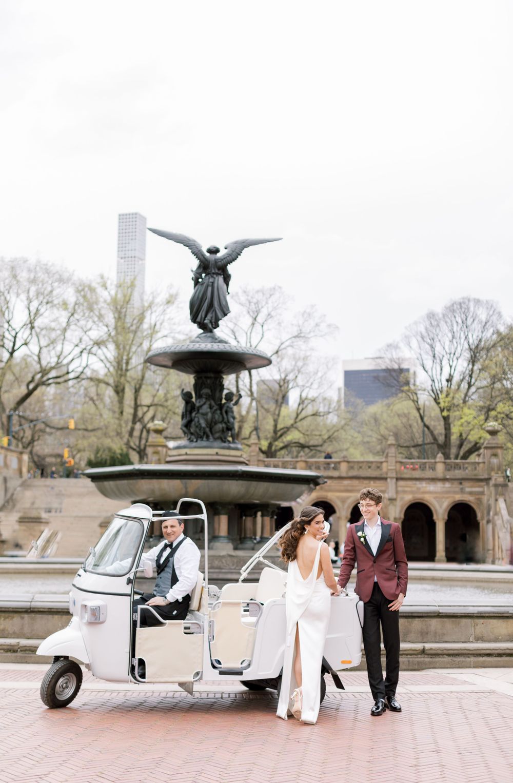 A wedding couple poses with a vintage white tuk tuk near Bethesda Fountain in Central Park.