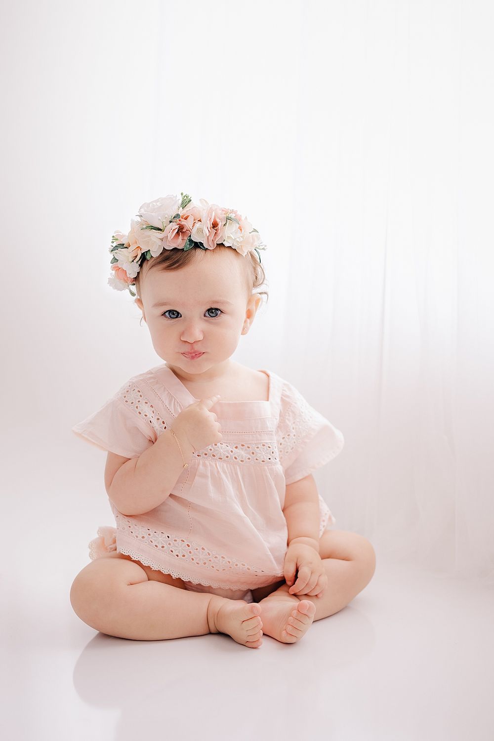 Baby in pink eyelet dress wearing floral crown against white backdrop.