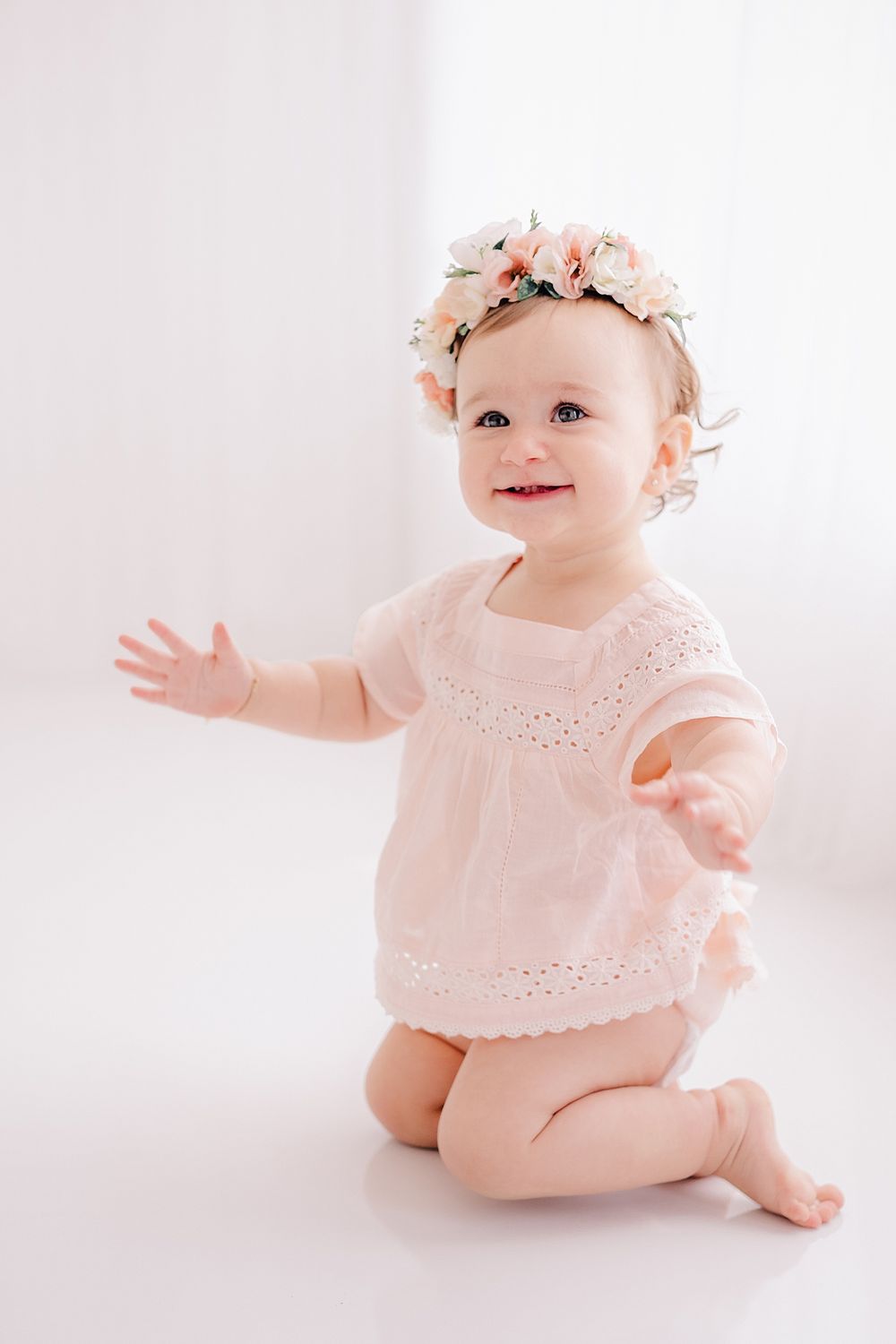 Baby in pink dress with floral crown reaching out against white background.