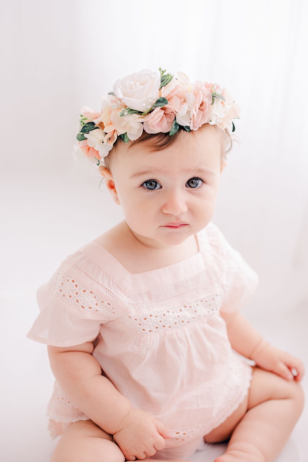 Close up portrait of baby wearing pink dress and peach floral crown against white backdrop.