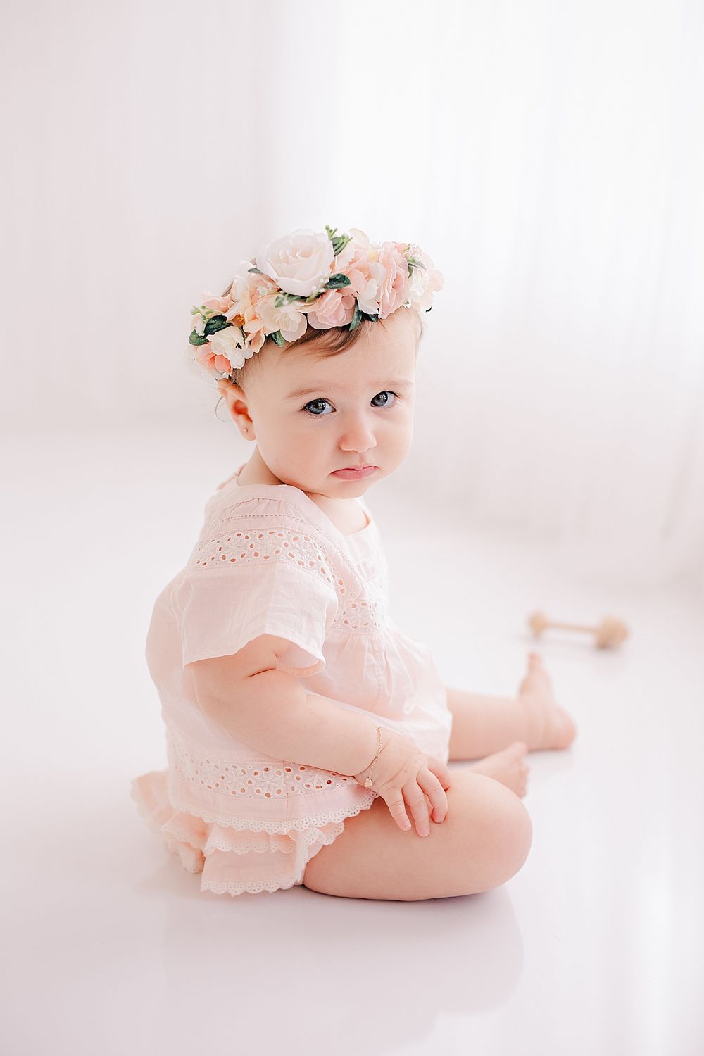 A baby wearing a pink lace romper and floral crown sits on a white background.