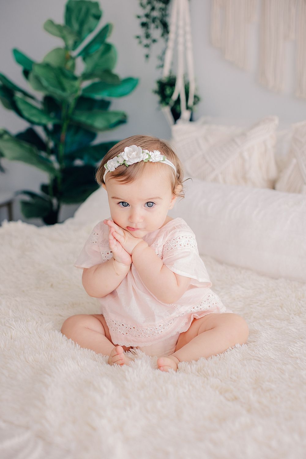 A baby in pink lace dress sits on white bedding with green plants in background.