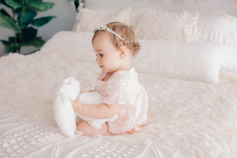 A baby in pink dress plays with a white plush bunny on textured white bedding.