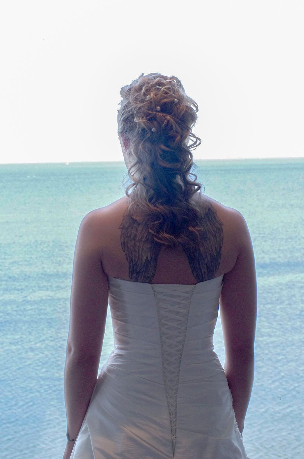 Back view of a bride in a white corset dress looking out at turquoise ocean waters.