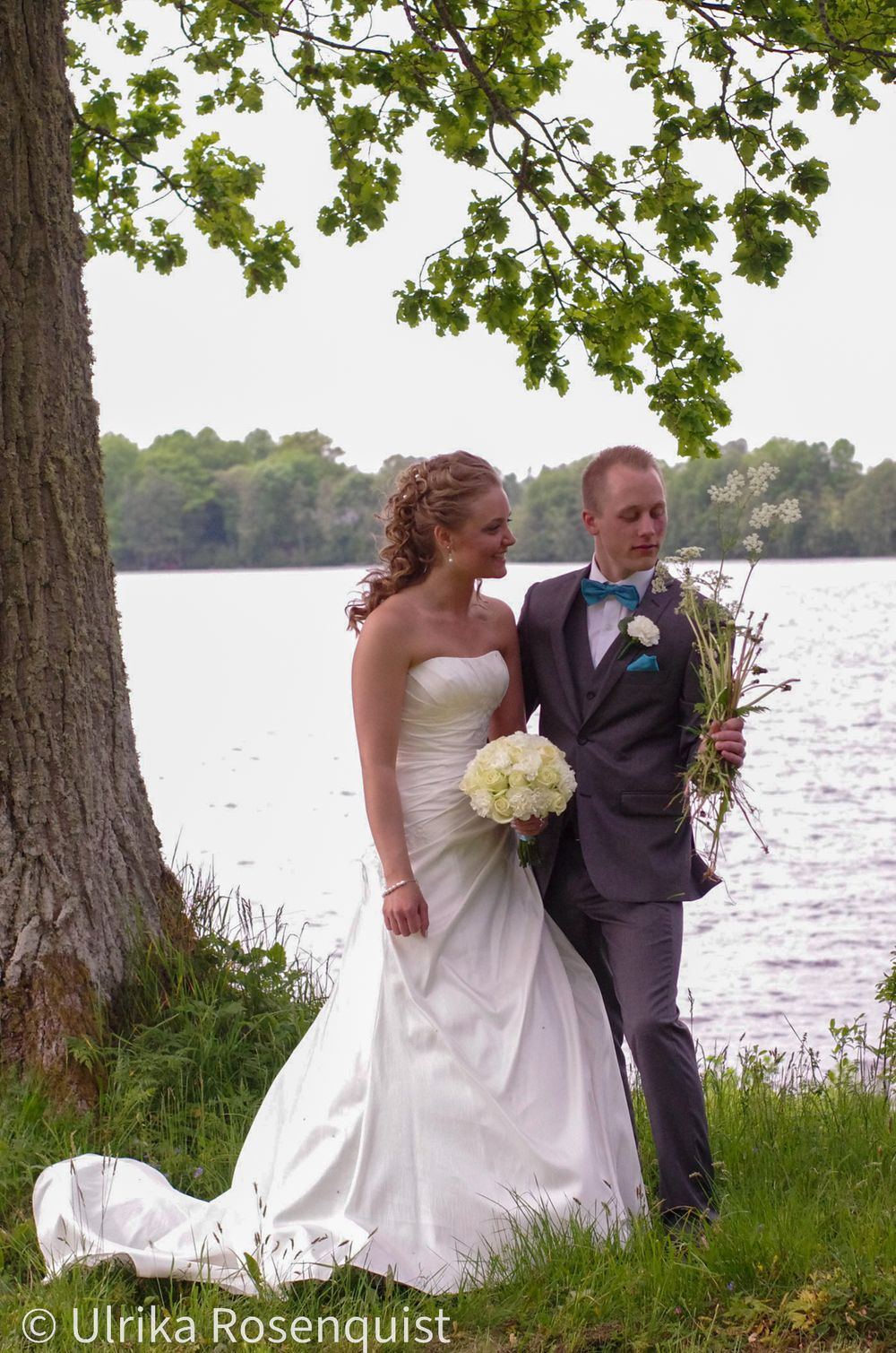 Newlywed couple posing by a lake under an oak tree on their wedding day.