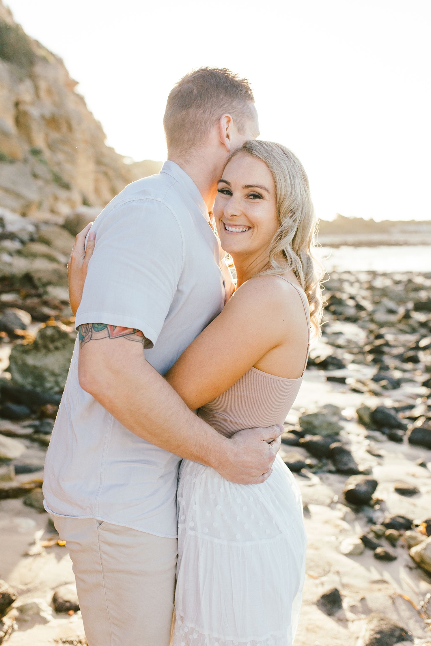 A couple embraces on a rocky beach with coastal cliffs and golden sunlight in the background.