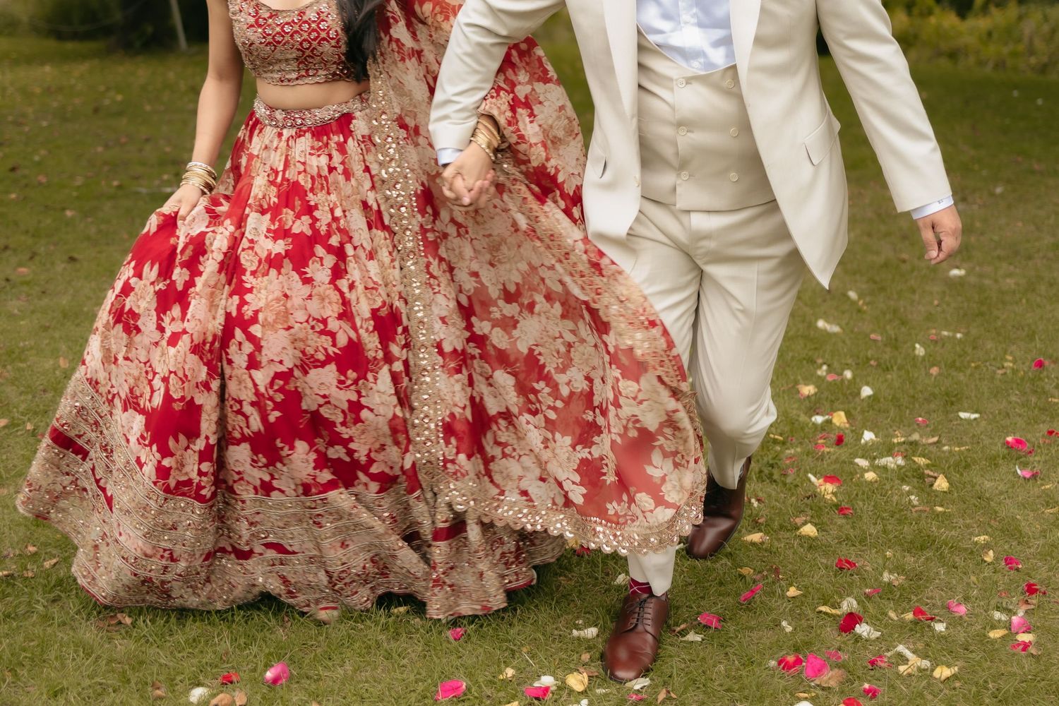 A red and beige floral wedding dress with scattered rose petals on grass and a cream-colored suit in the background.