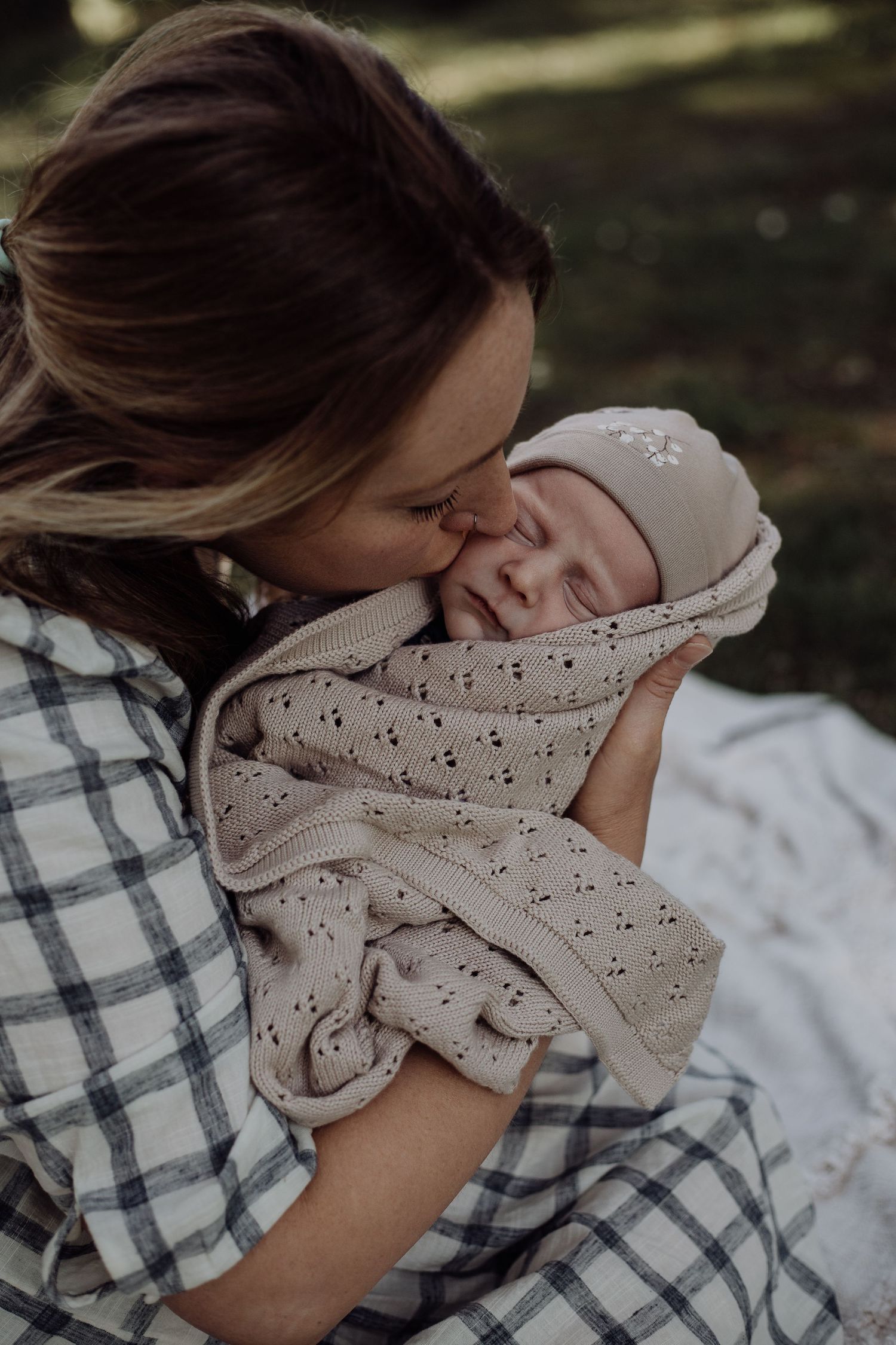 Mother in plaid shirt tenderly kissing newborn baby wrapped in patterned blanket outdoors.