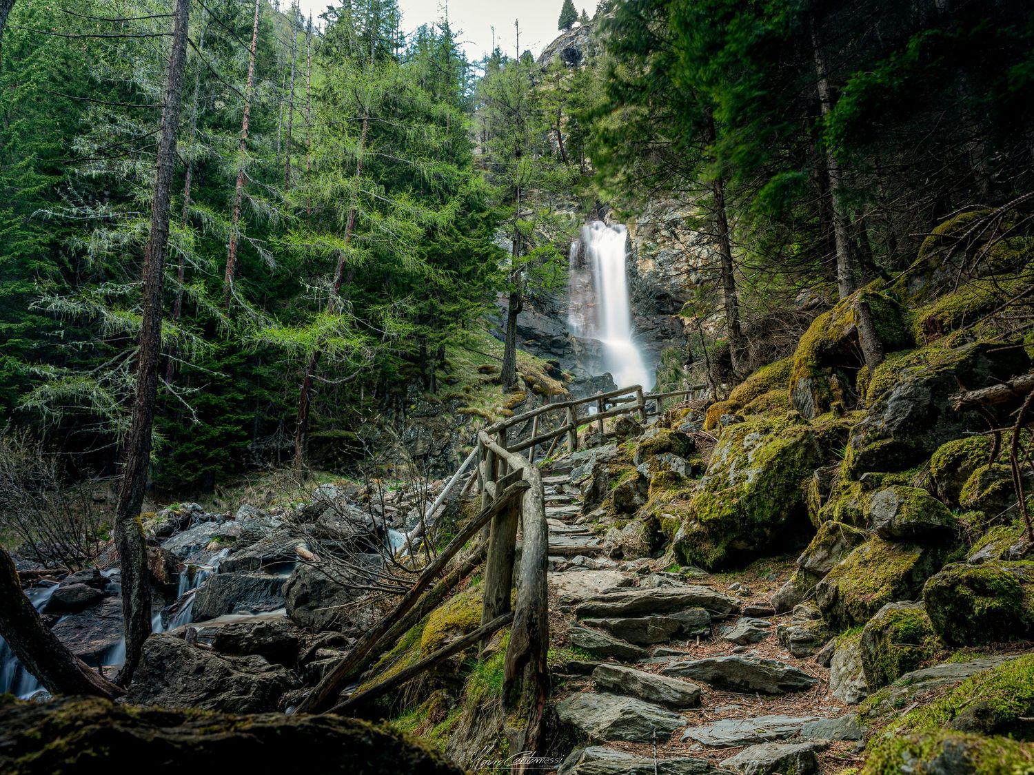 A scenic waterfall cascades down moss-covered rocks with wooden steps leading up the mountainside.
