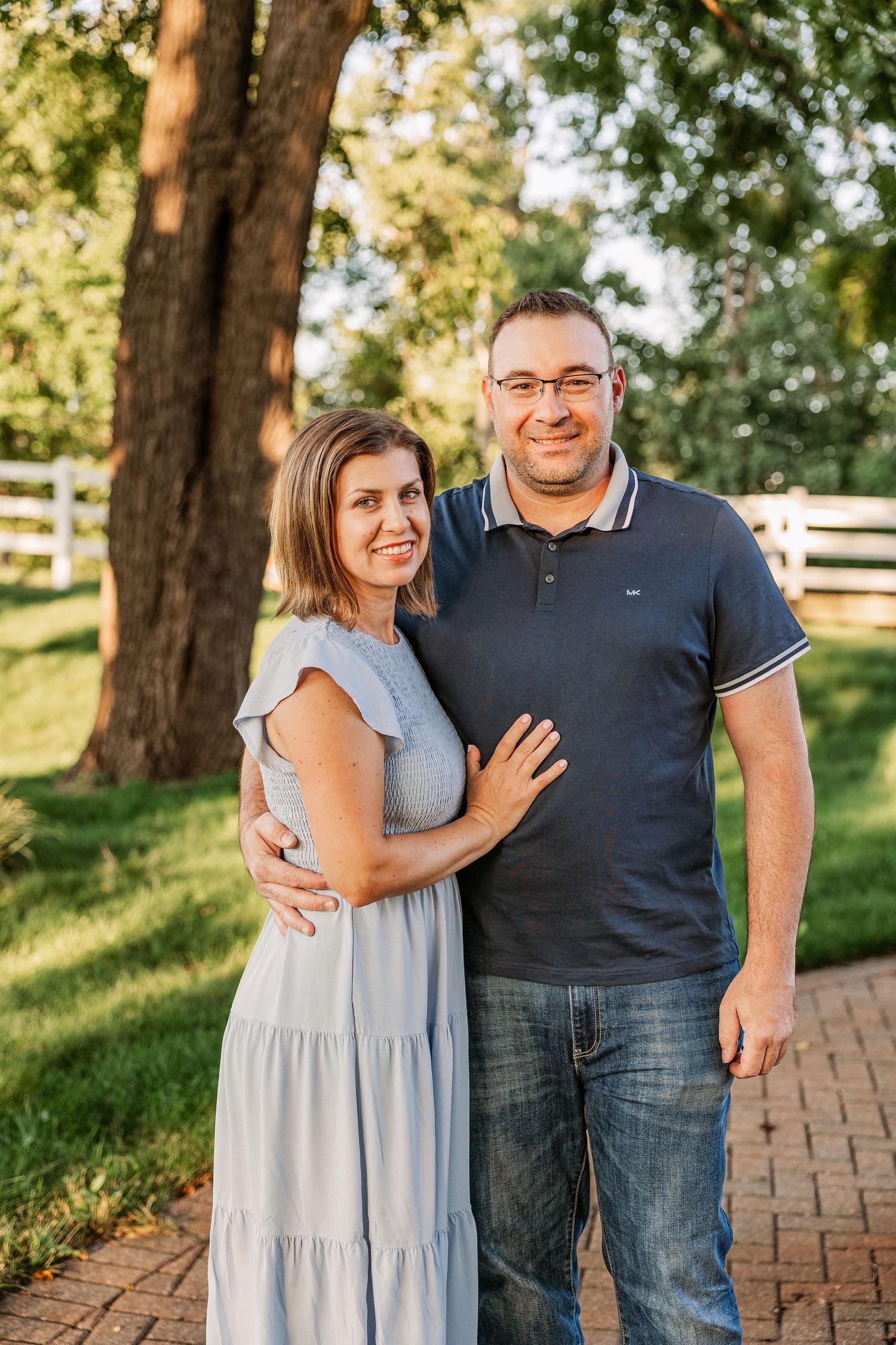 A couple poses together outdoors on a brick pathway with white fence and trees in the background.