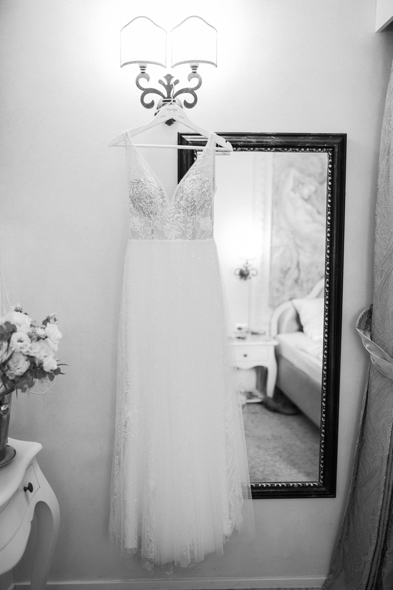 A white lace wedding dress hangs elegantly against a wall with decorative sconces.