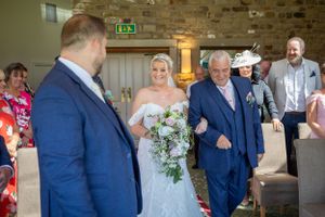 Bride walks down the aisle with her father toward the groom in a rustic stone venue.