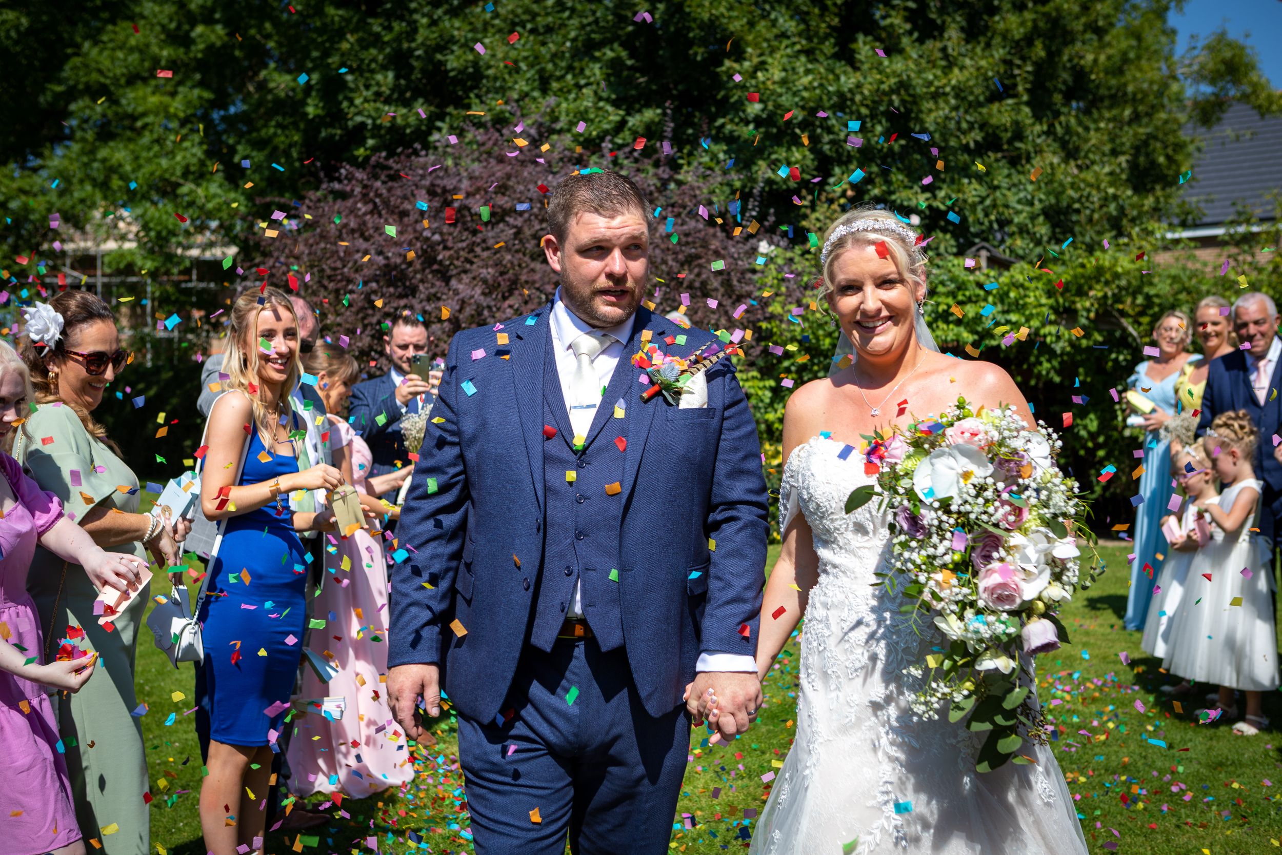 Bride and groom walk through colorful confetti shower surrounded by cheering wedding guests outdoors.