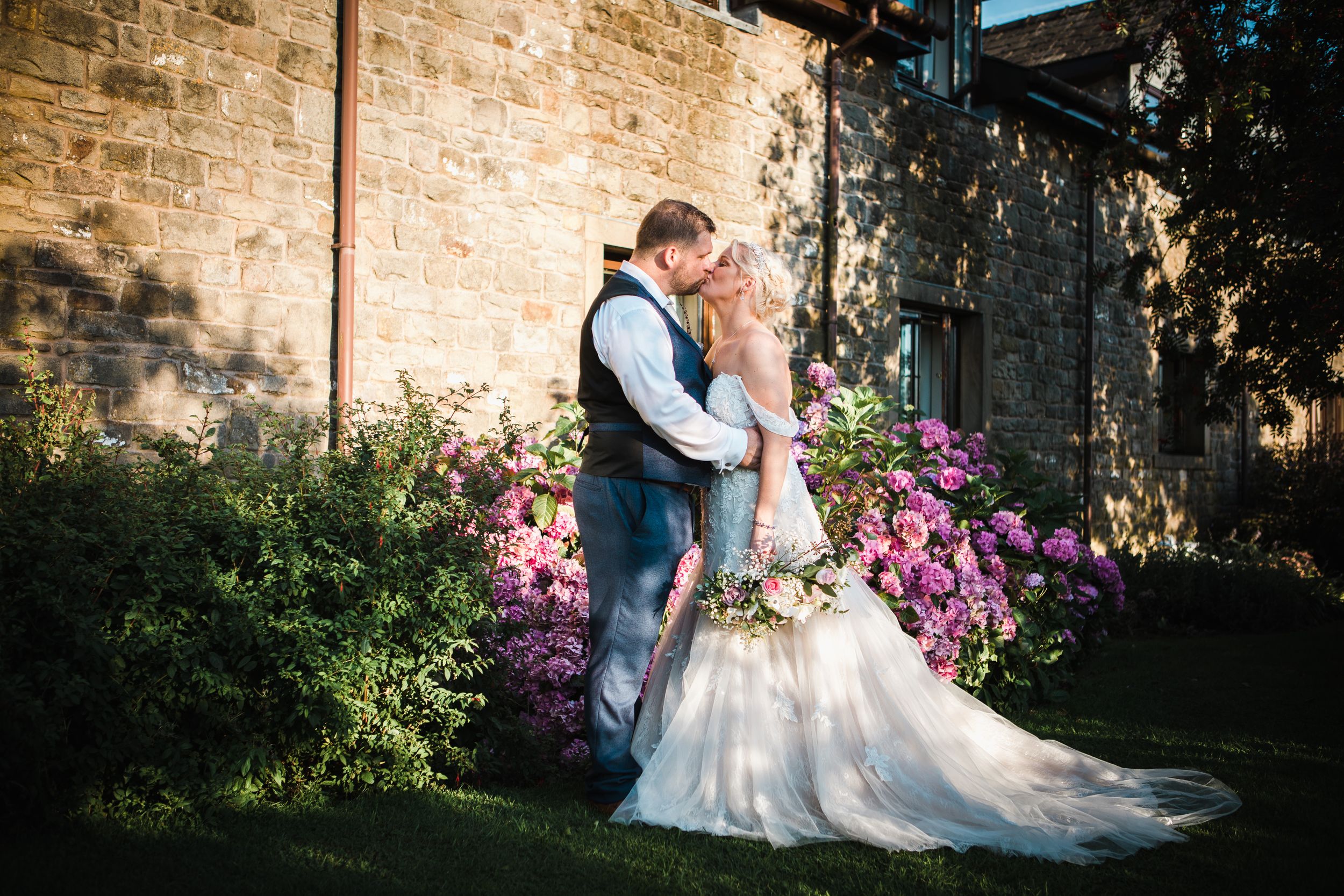 Bride and groom share a tender kiss surrounded by vibrant pink roses outside a stone building.