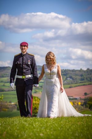 Military groom in red beret and bride in lace gown walk hand in hand over scenic green hills.