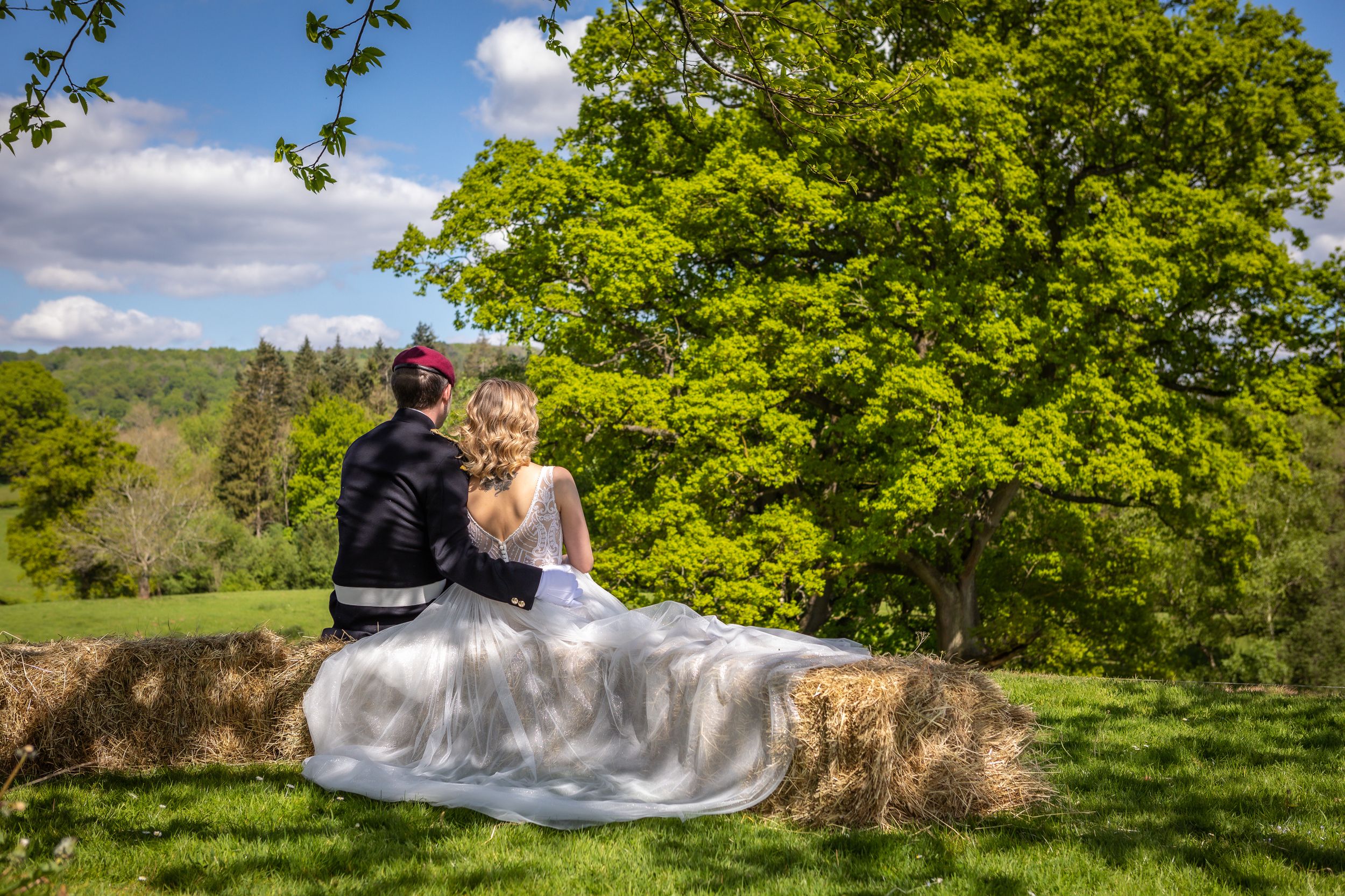 Romantic bride and groom sit together on a stone wall surrounded by lush green trees on their wedding day.