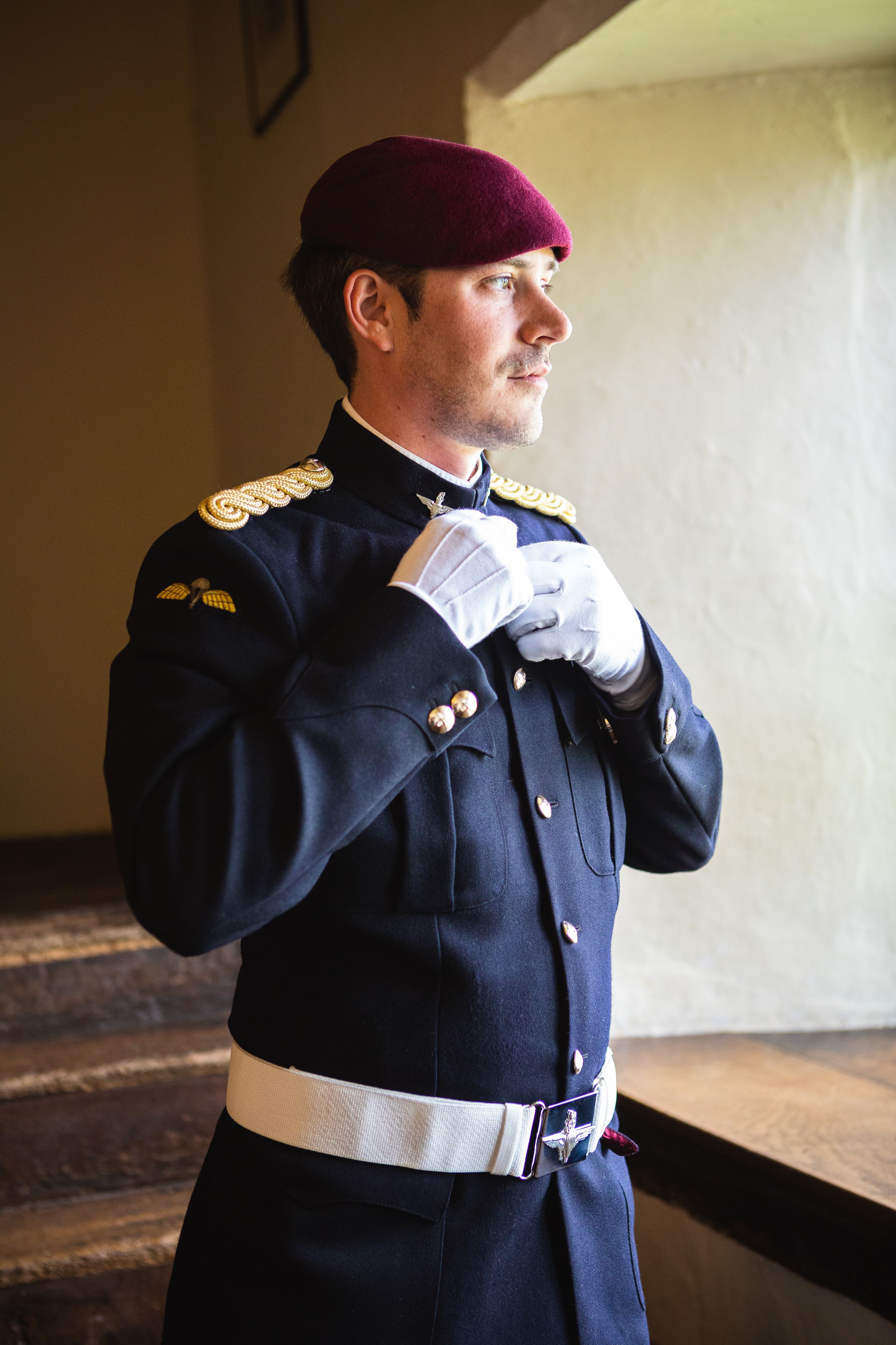 Groom in military dress uniform with maroon beret adjusting his collar before wedding ceremony.