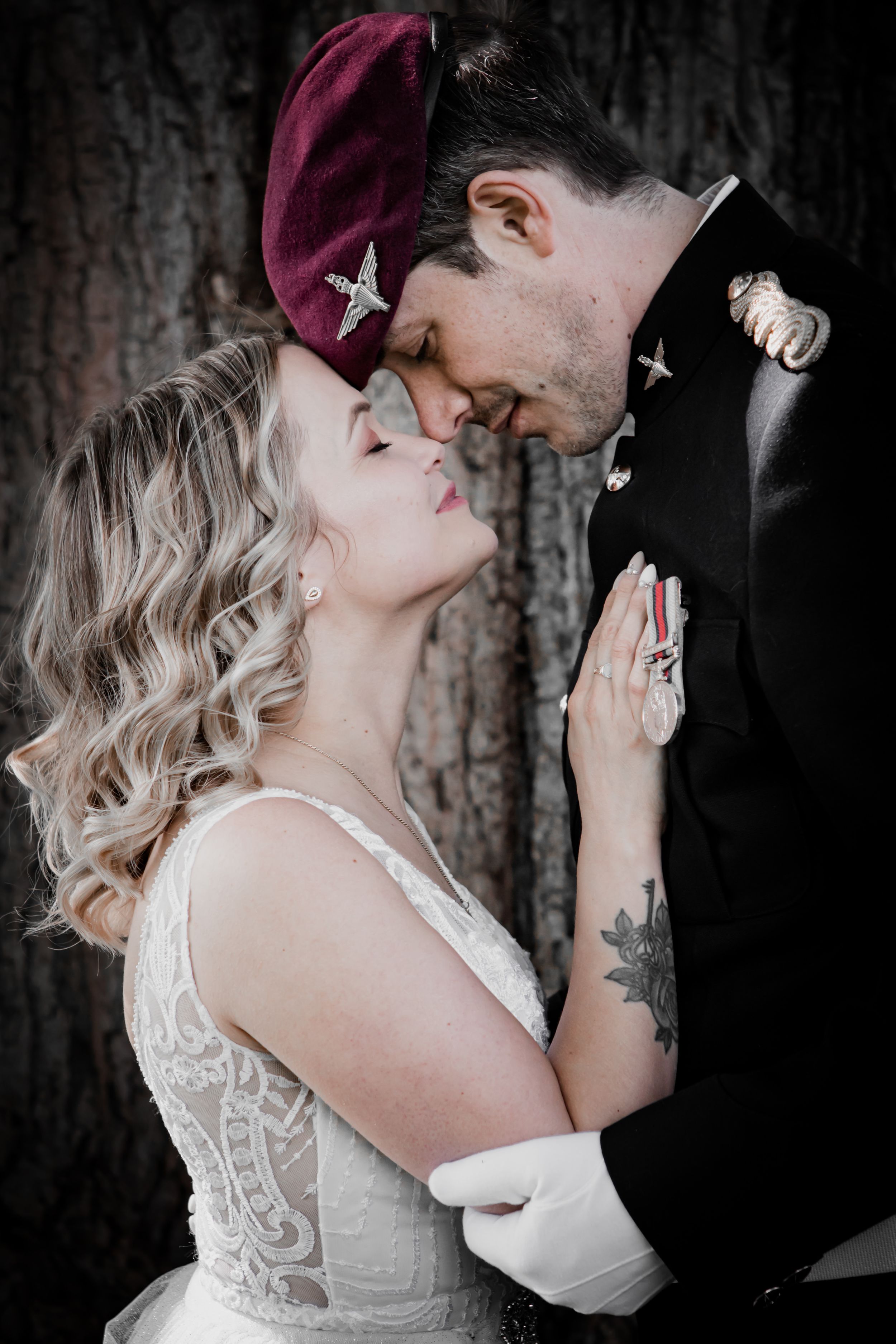 Bride and military groom share an intimate moment, touching foreheads tenderly against a tree backdrop.