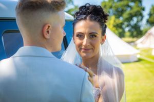 Beautiful bride in veil gazes lovingly at groom near a vintage blue van at an outdoor summer wedding.